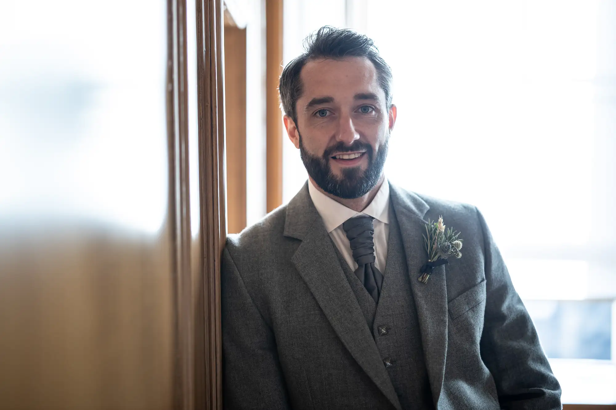 Man in a gray suit with a boutonniere smiling, standing by a window indoors, sunlight filtering in.