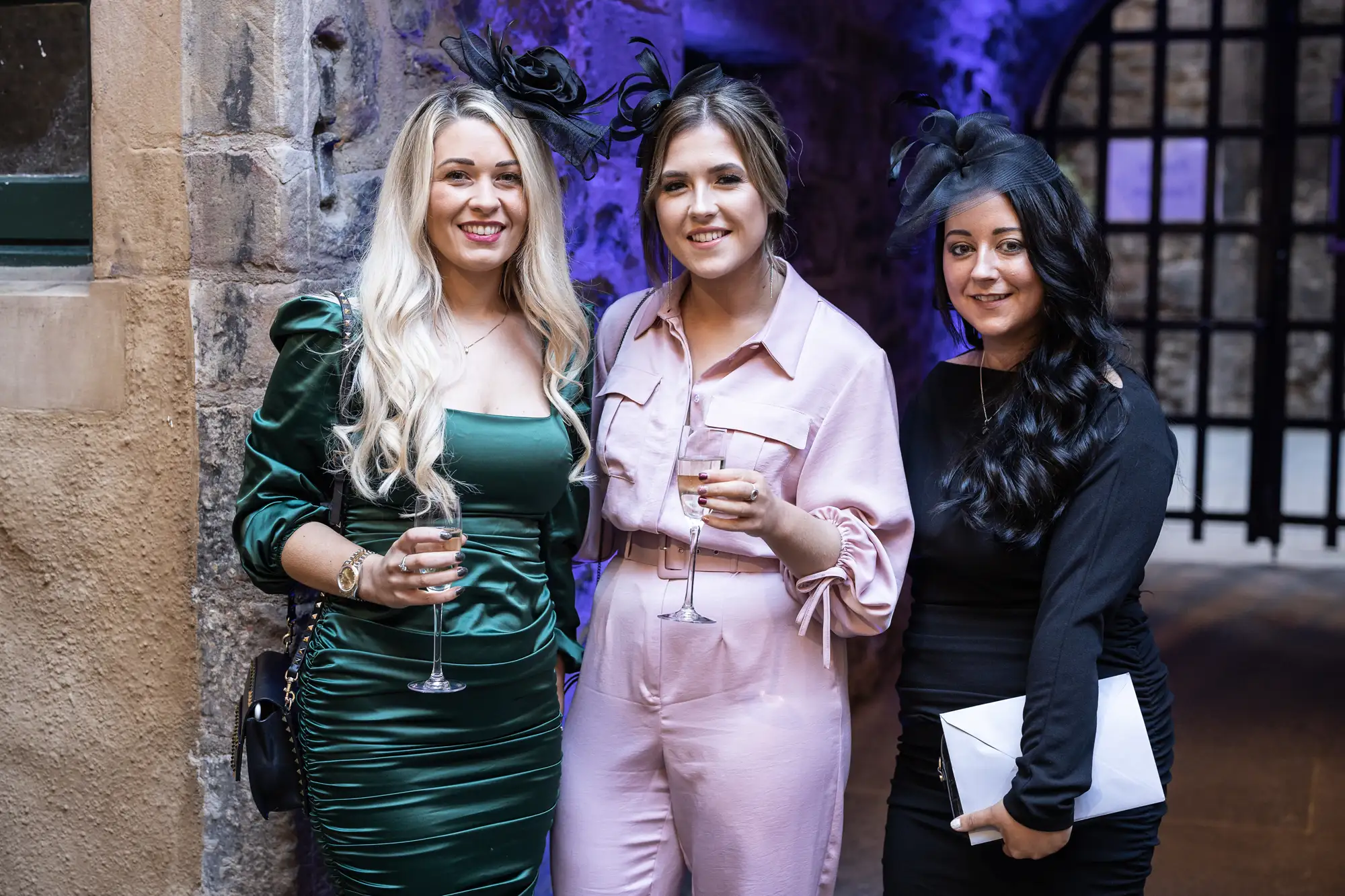 Three women smiling at a social event, each holding a glass of wine, dressed in formal attire in a rustic setting.