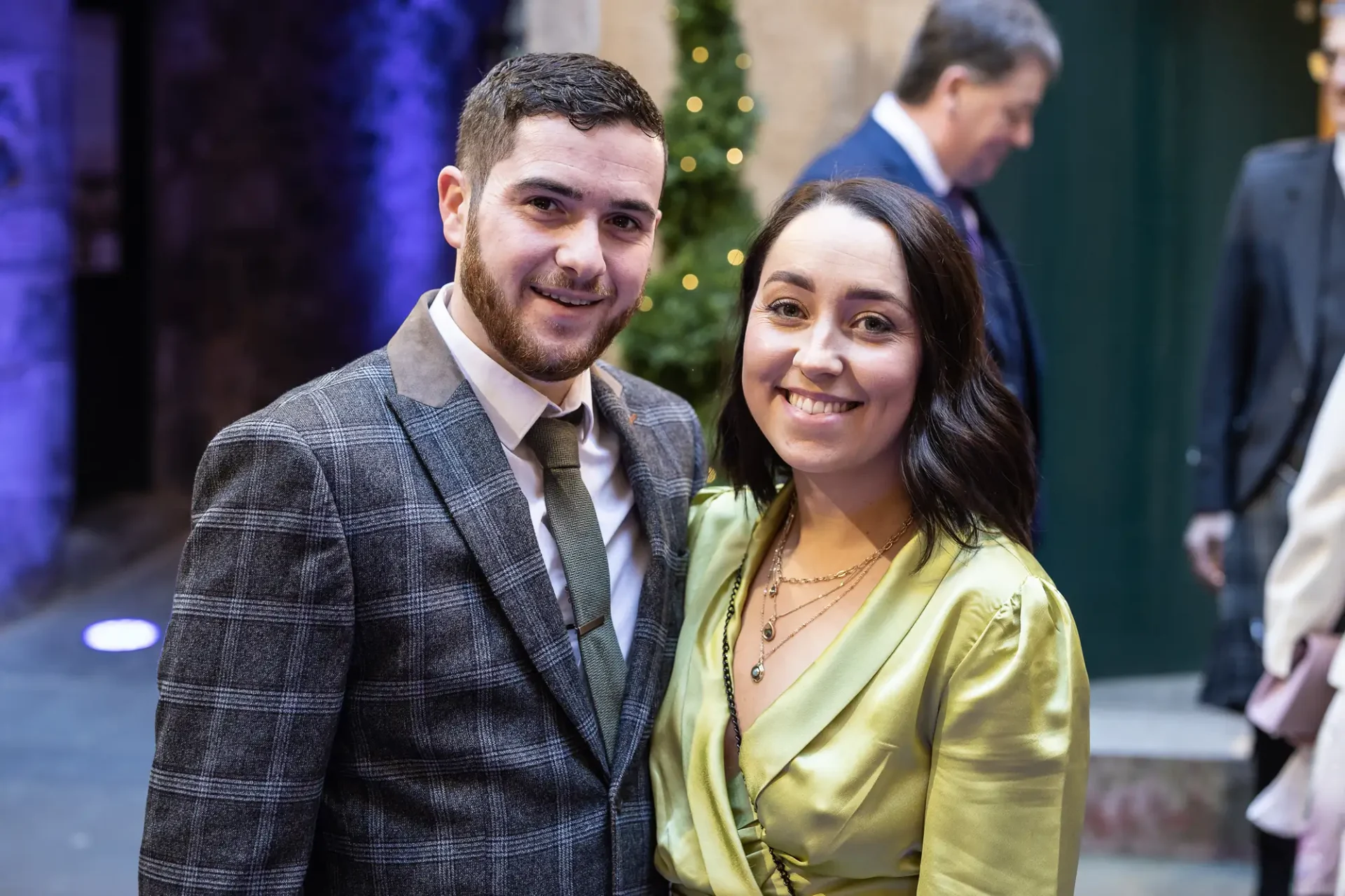 A smiling couple, the man in a plaid suit and the woman in a yellow dress, posing together at a formal event with festive decorations in the background.