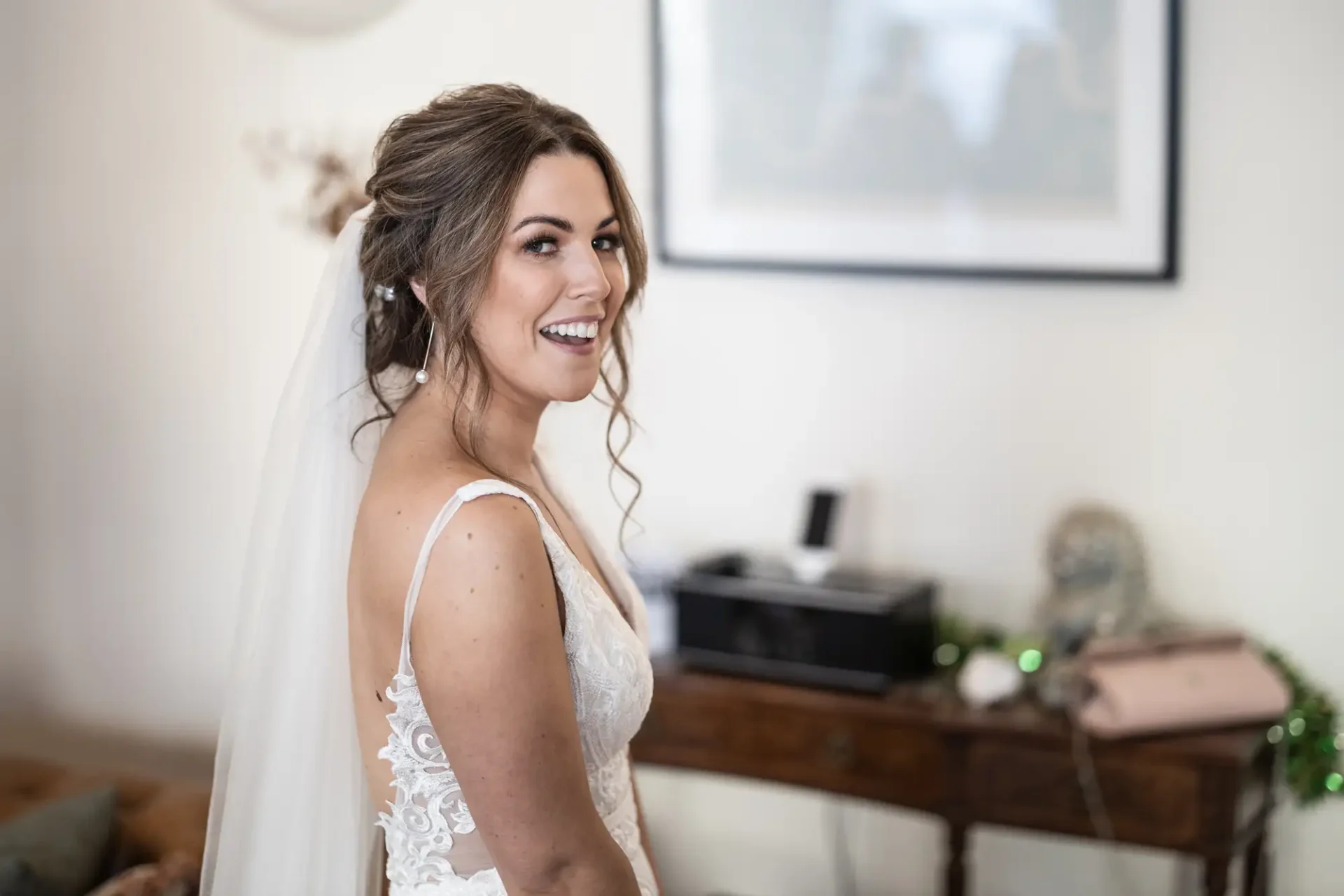 A smiling bride in a lace wedding dress and veil, looking over her shoulder in a tastefully decorated room.