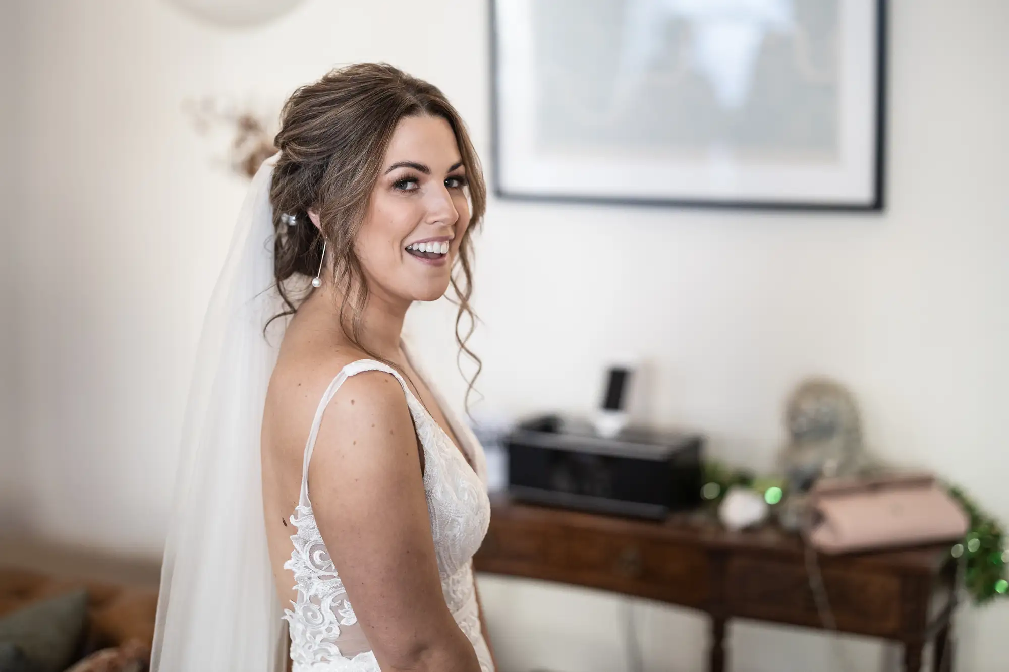 A smiling bride in a lace wedding dress and veil, looking over her shoulder in a tastefully decorated room.