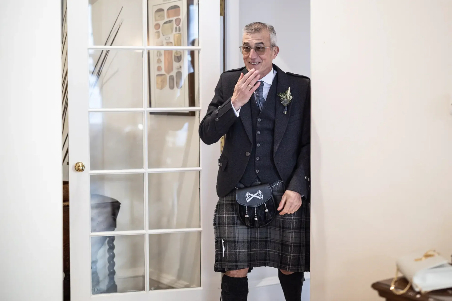 Man in traditional scottish attire with a kilt, smiling and waving in a room with a glass door and wall display.