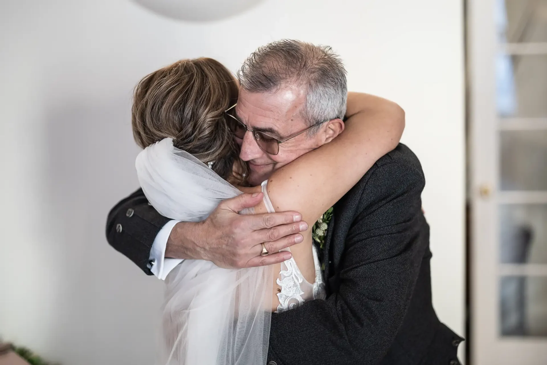 An older man in a suit hugs a younger woman in a white wedding dress, both smiling during an emotional embrace.