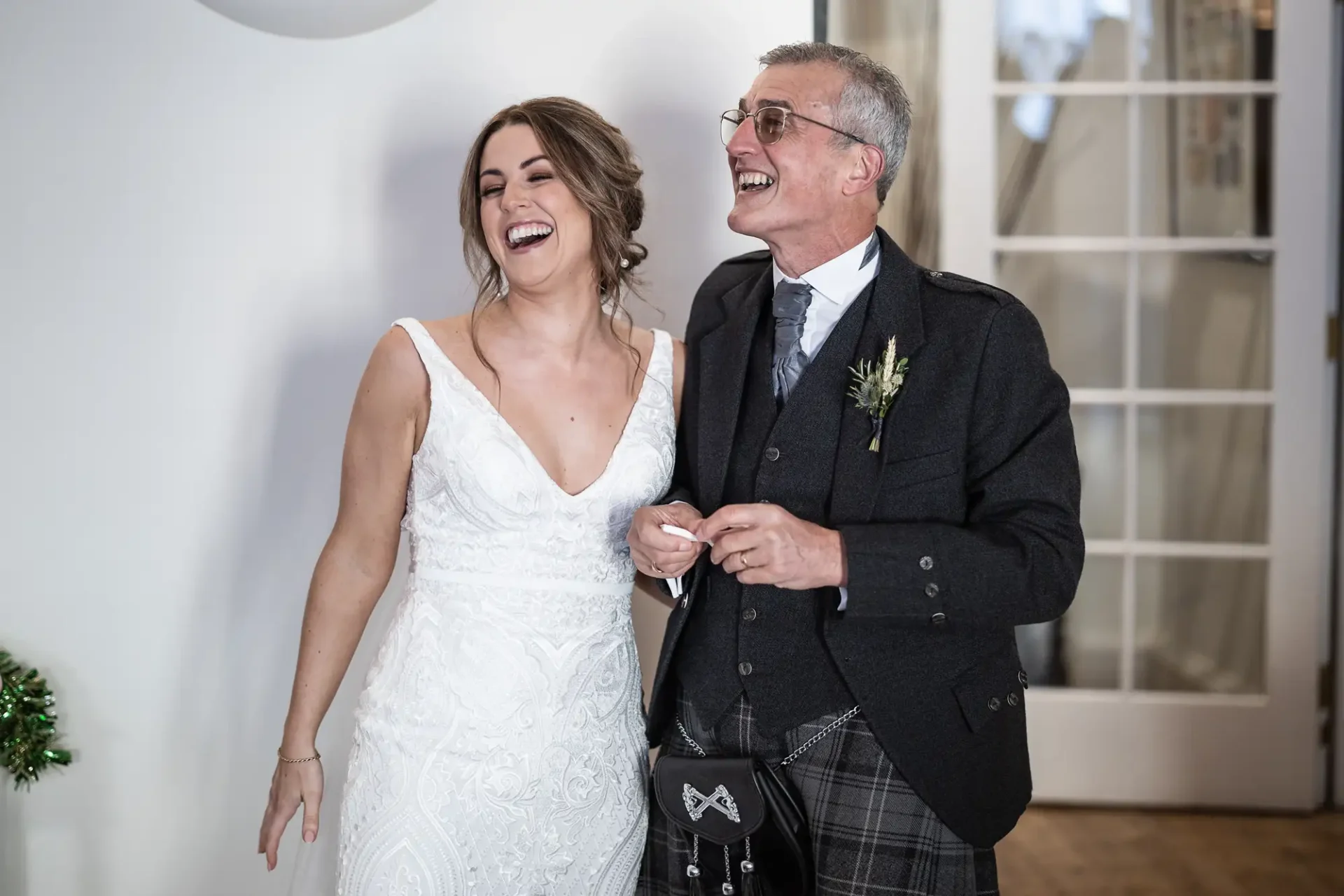 A bride in a white dress laughs joyfully next to an older man in a kilt and tweed jacket, both holding hands at a wedding.