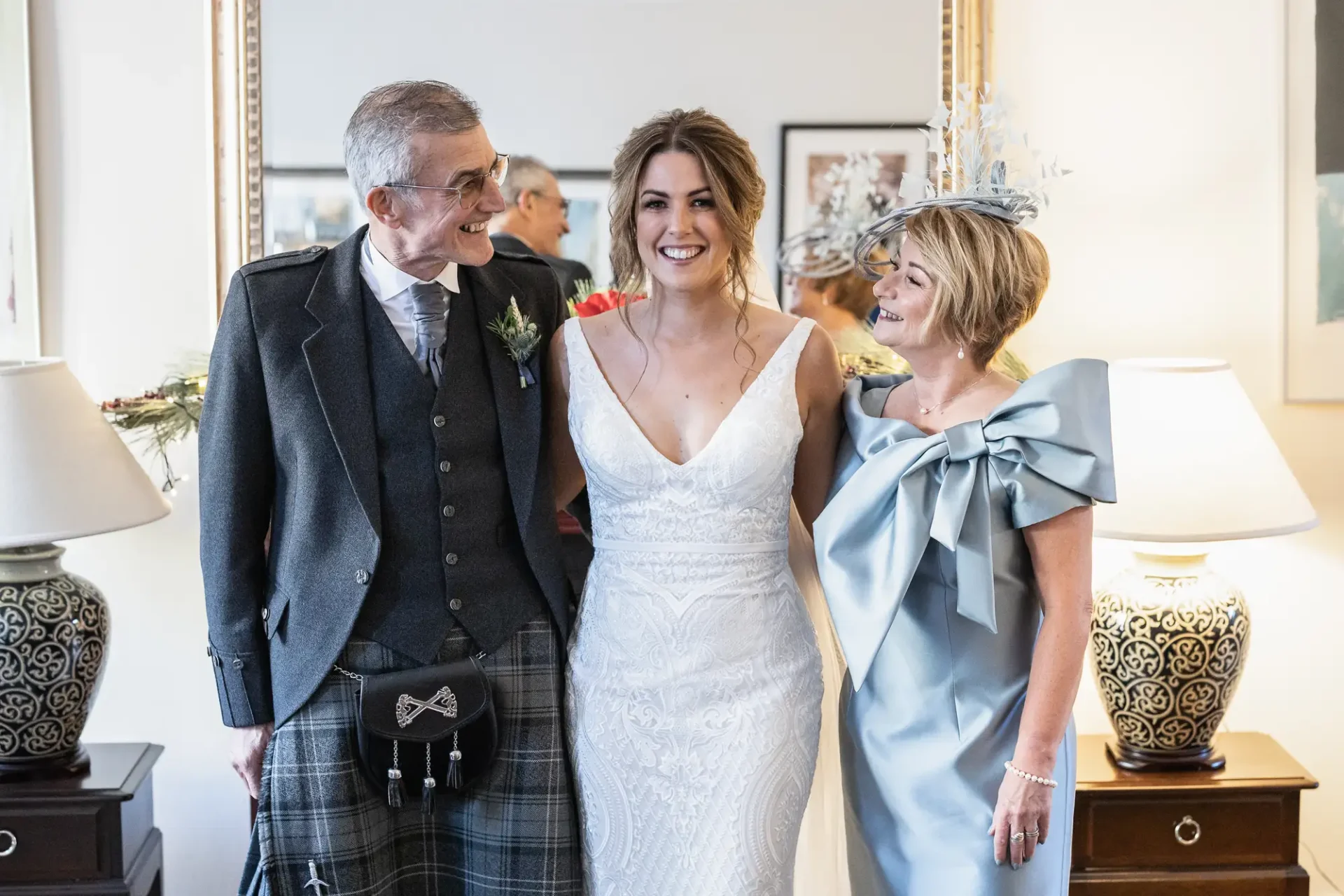 A bride in a white dress smiling with an older man in a kilt and a woman in a blue dress and hat indoors.