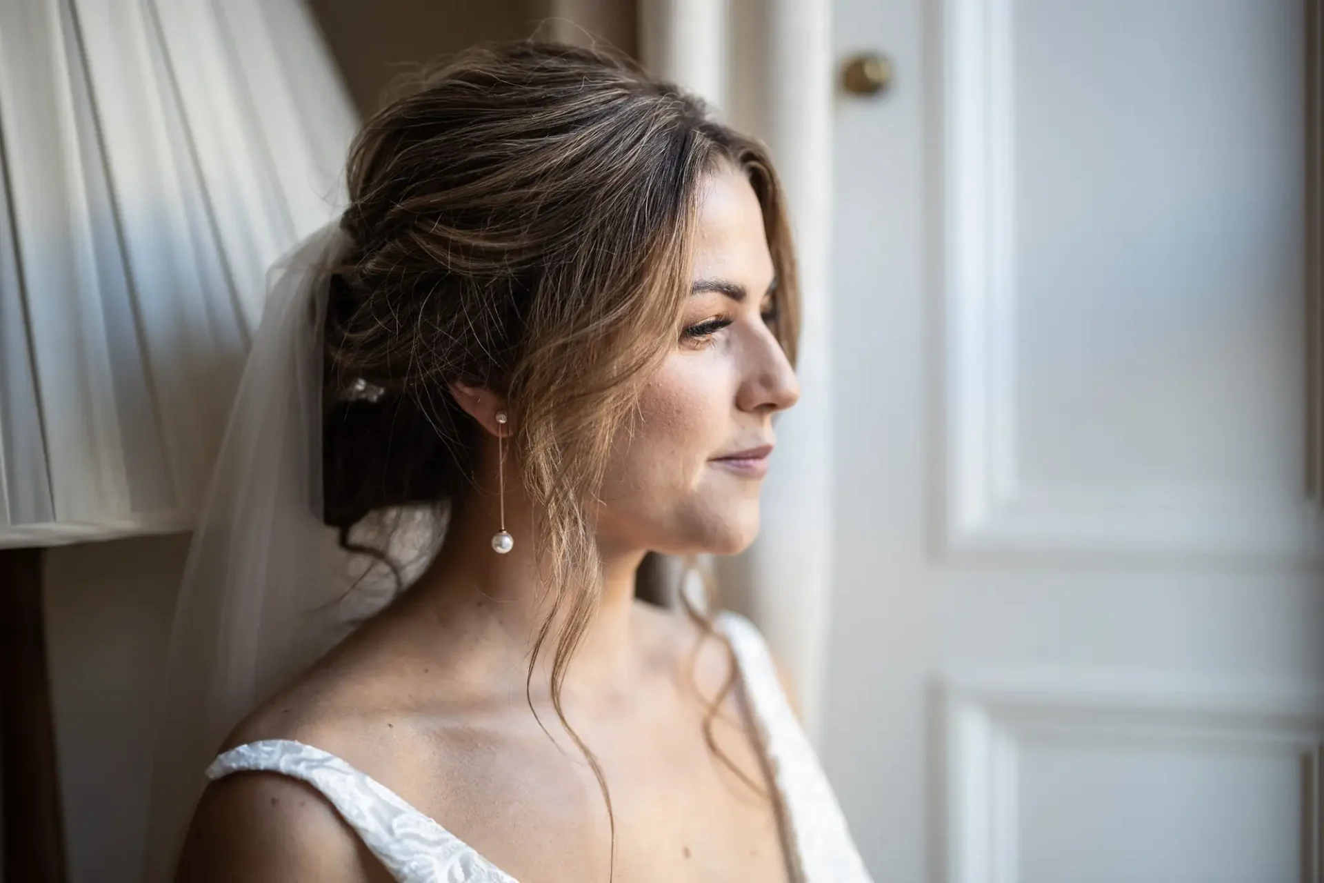 A bride in an off-shoulder wedding dress looks contemplative by a window, showcasing a styled updo with a veil and pearl earrings.