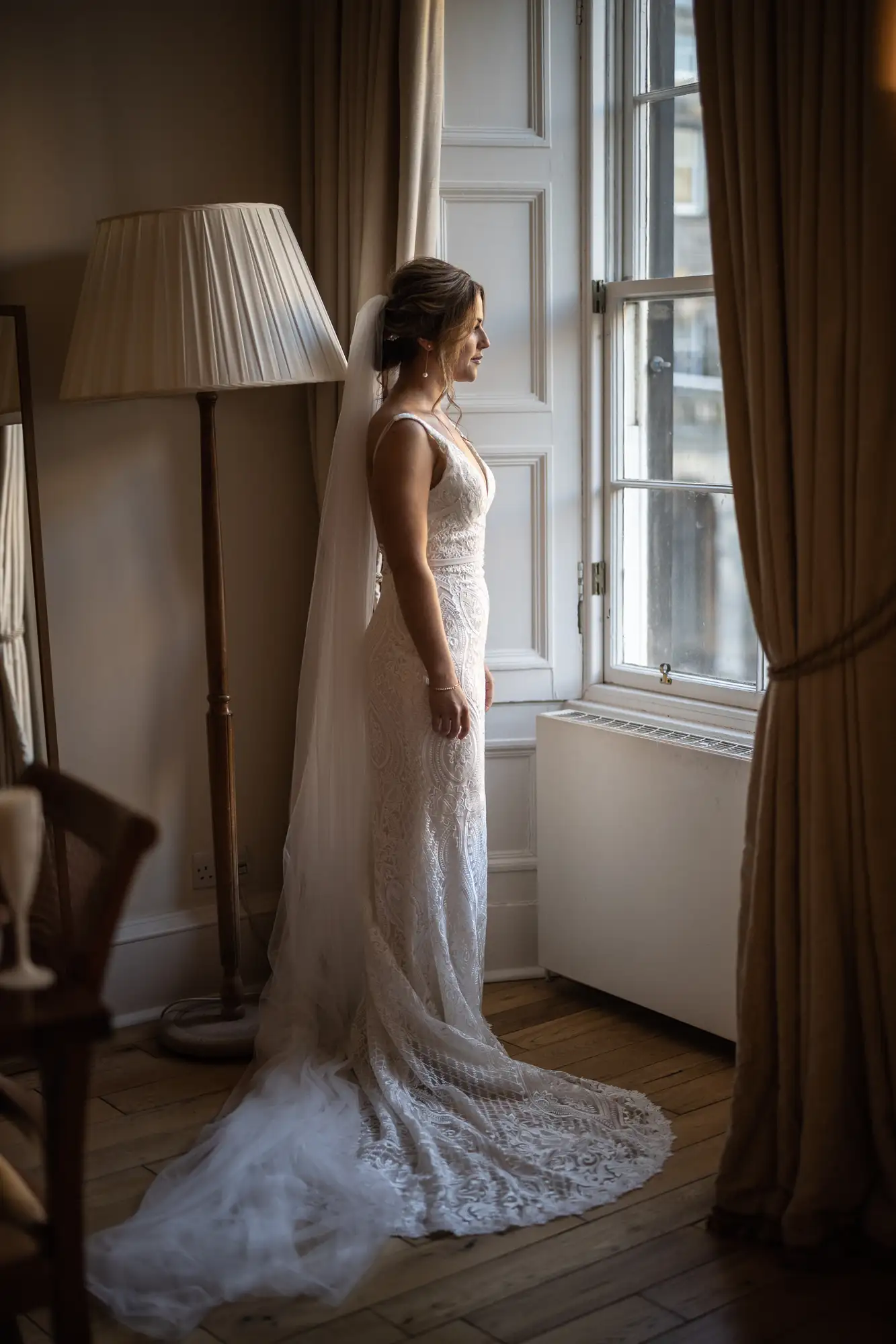 A bride in a lace gown and veil stands by a window, looking out, in a warmly lit room with elegant furniture.