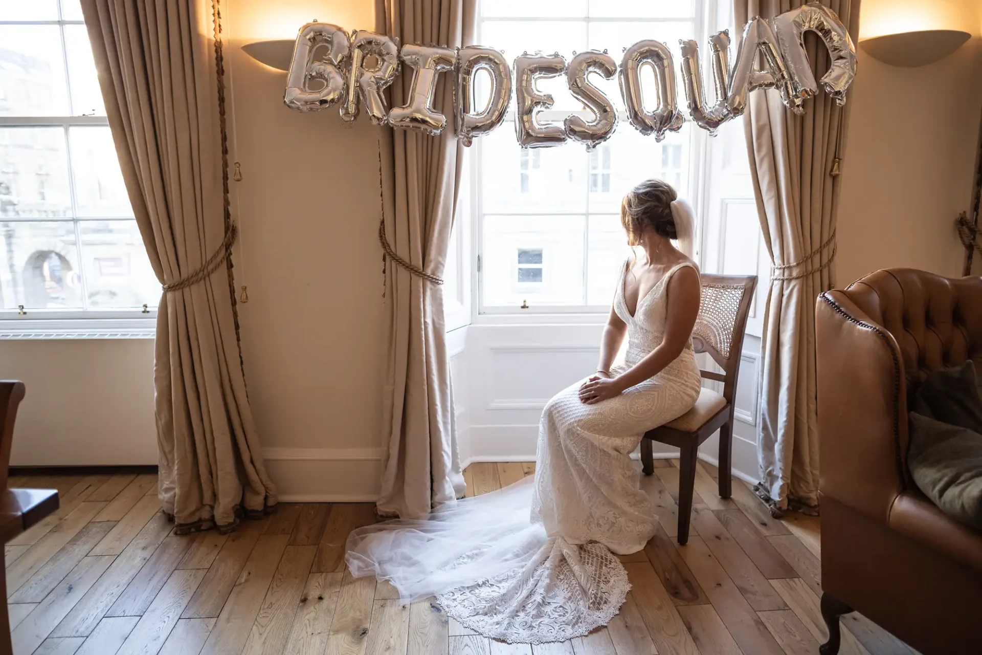 A bride in a long white dress sits pensively on a chair under a "bride squad" balloon sign, with large windows on either side in a well-lit room.