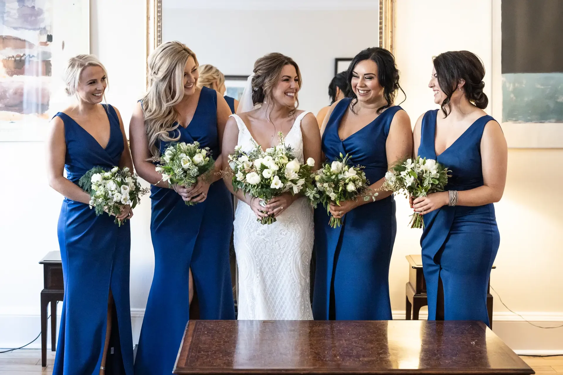 Five women in blue dresses, holding bouquets and smiling joyously, stand beside a bride in a white gown.