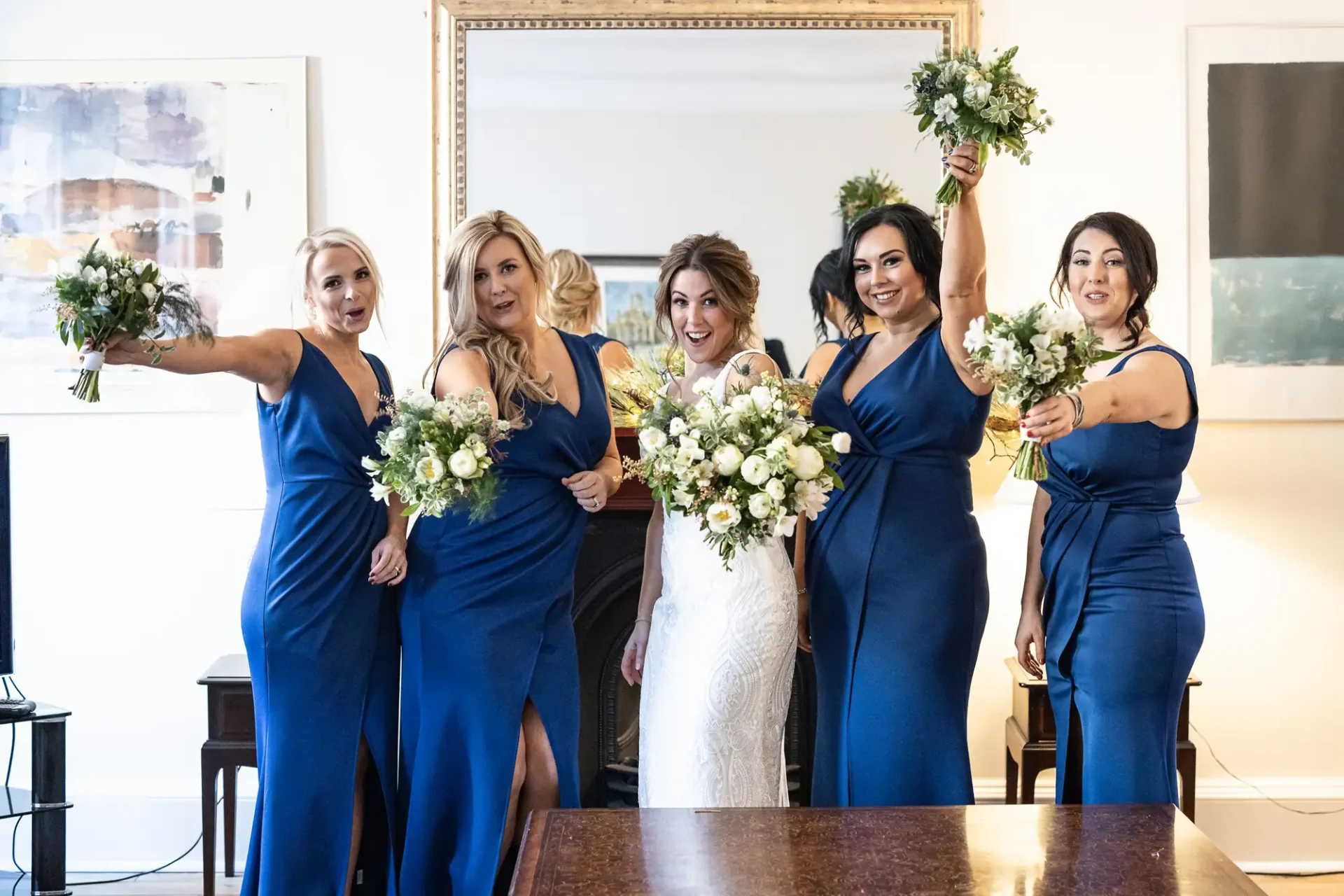 Bride with four bridesmaids in blue dresses, holding bouquets and posing excitedly in an elegantly decorated room.