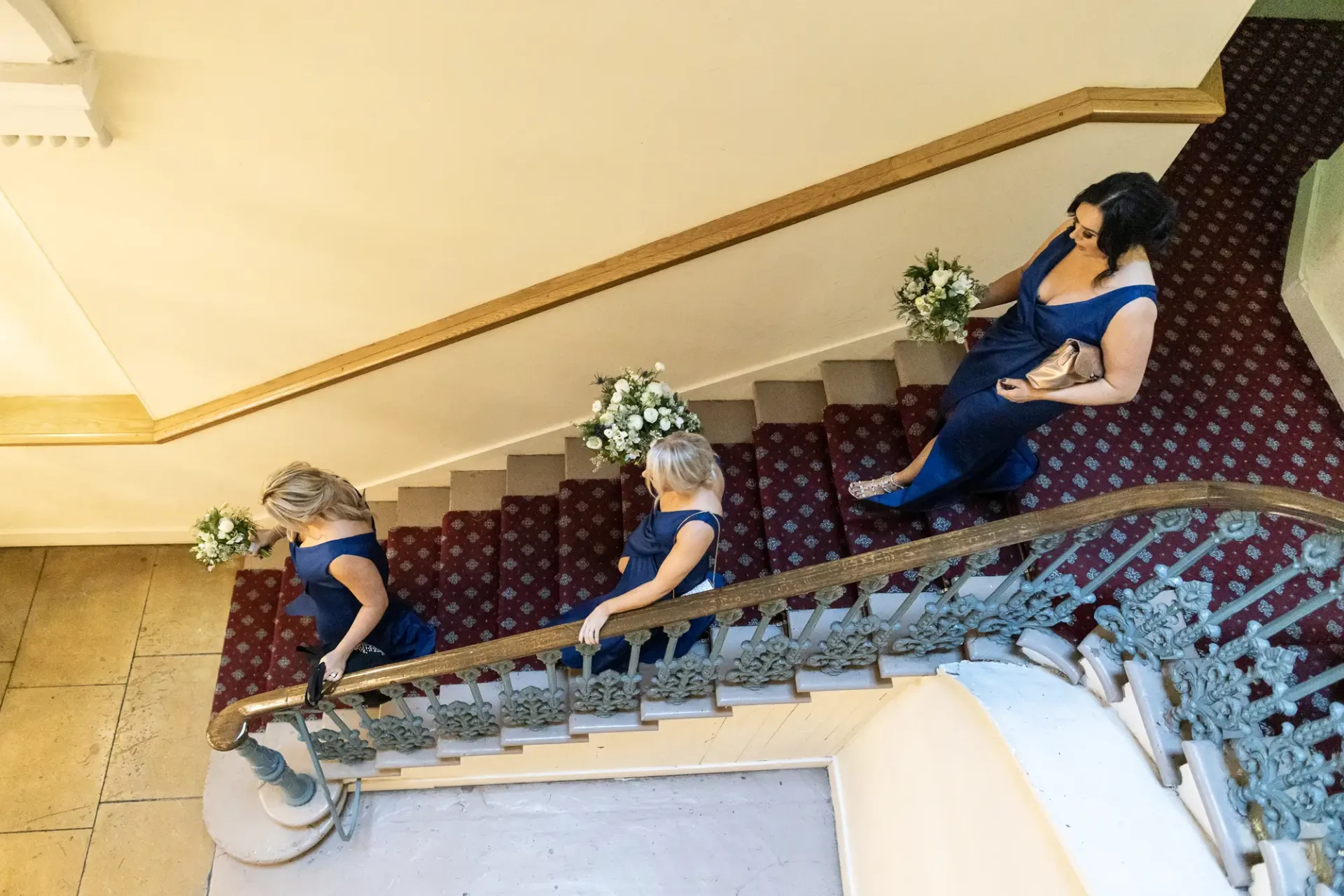 Two women and a girl in blue dresses descend a grand staircase, holding bouquets; another woman sits on the steps.