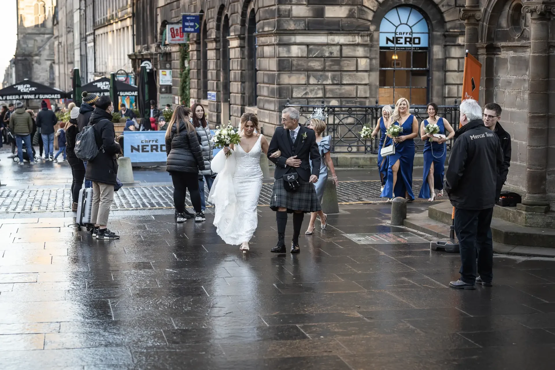 A bride in a white dress, escorted by an older man, walks through a bustling city street with onlookers, some in kilts.