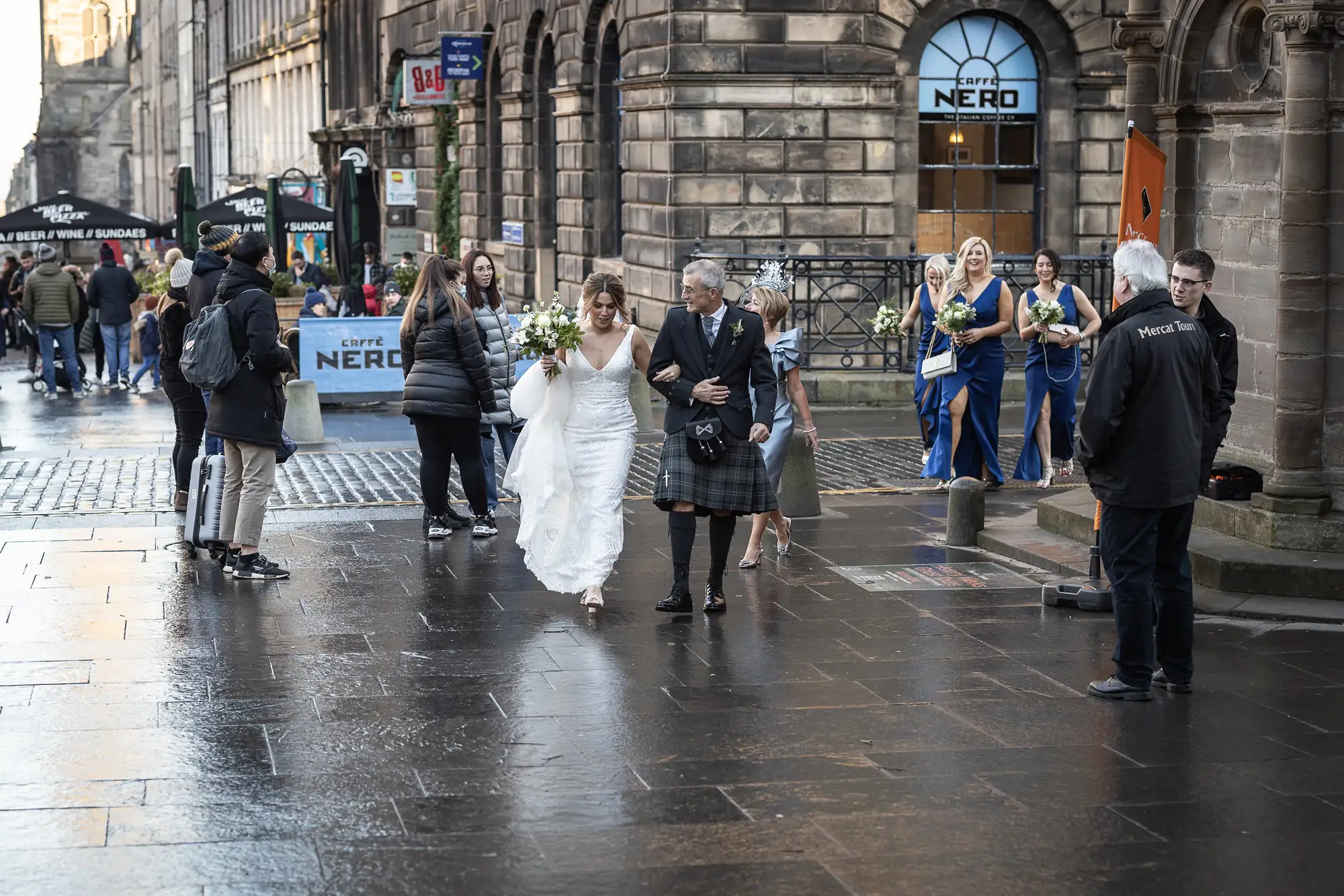 A bride in a white dress, escorted by an older man, walks through a bustling city street with onlookers, some in kilts.