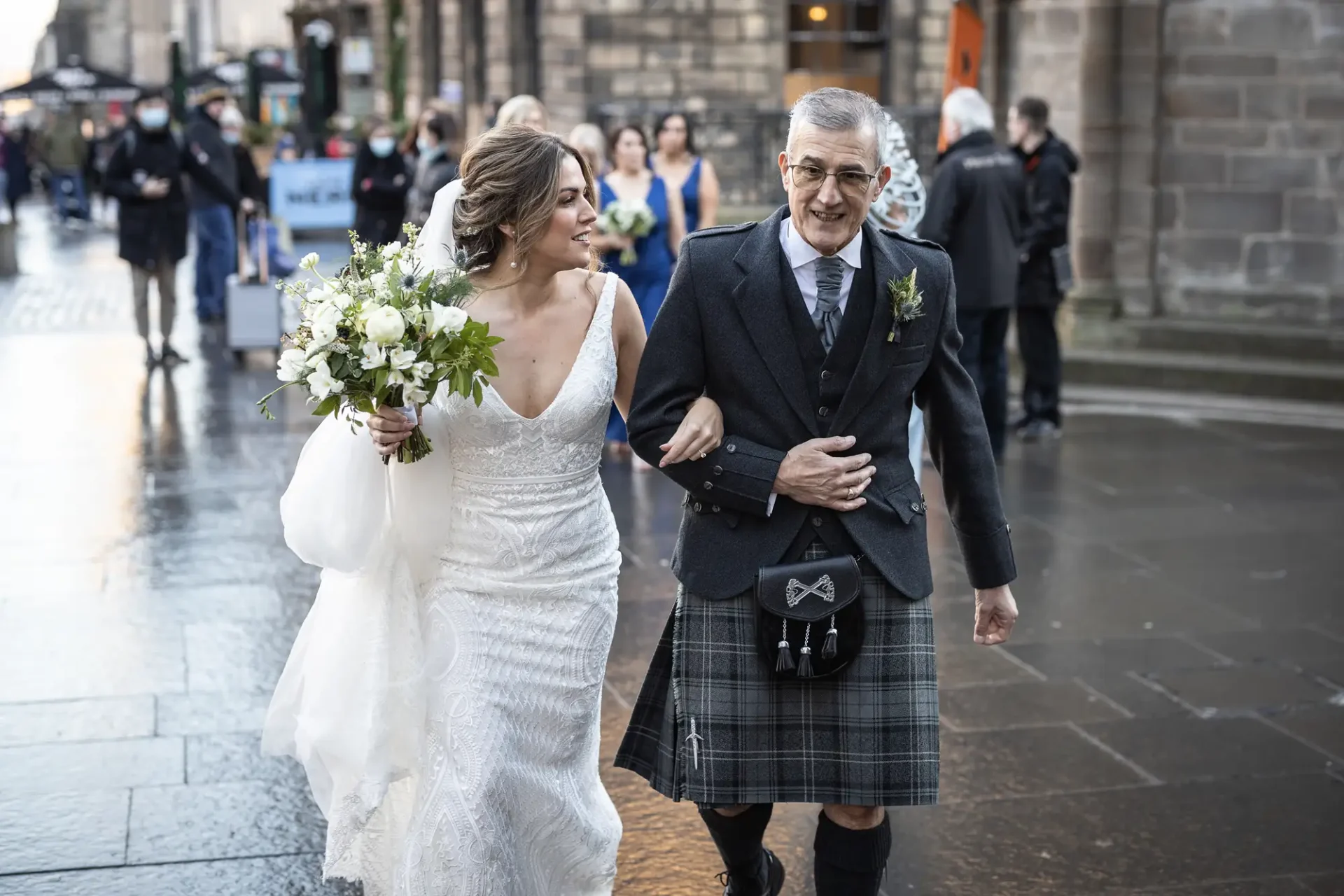 A bride in a white dress walks down a wet street with an older man in a kilt and sporran, both smiling joyfully.
