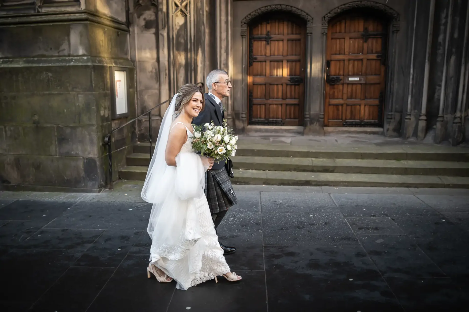 Bride in a white dress walking with an older man in a kilt outside a church with large wooden doors.