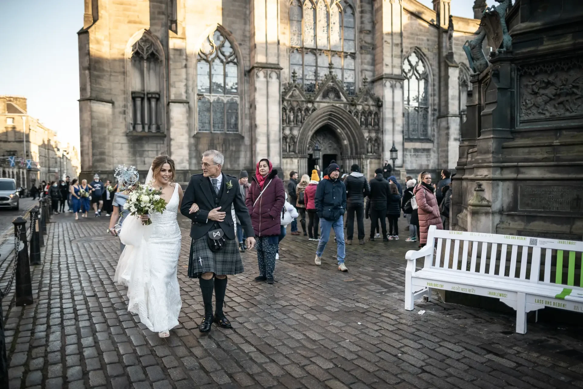 A bride and groom, dressed in traditional scottish attire, walk hand-in-hand outside a historic cathedral, with onlookers in the background.