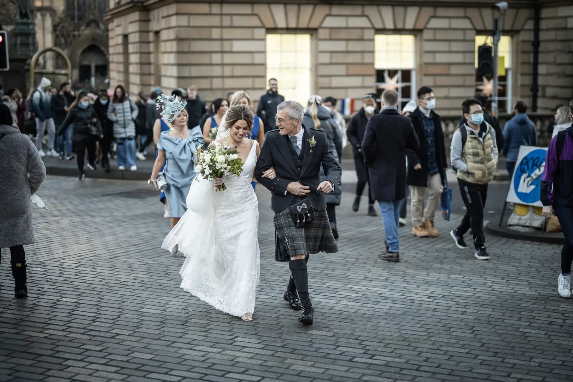 Bride in a white dress and an elderly man in a kilt walking joyfully across a busy street, surrounded by onlookers wearing masks.