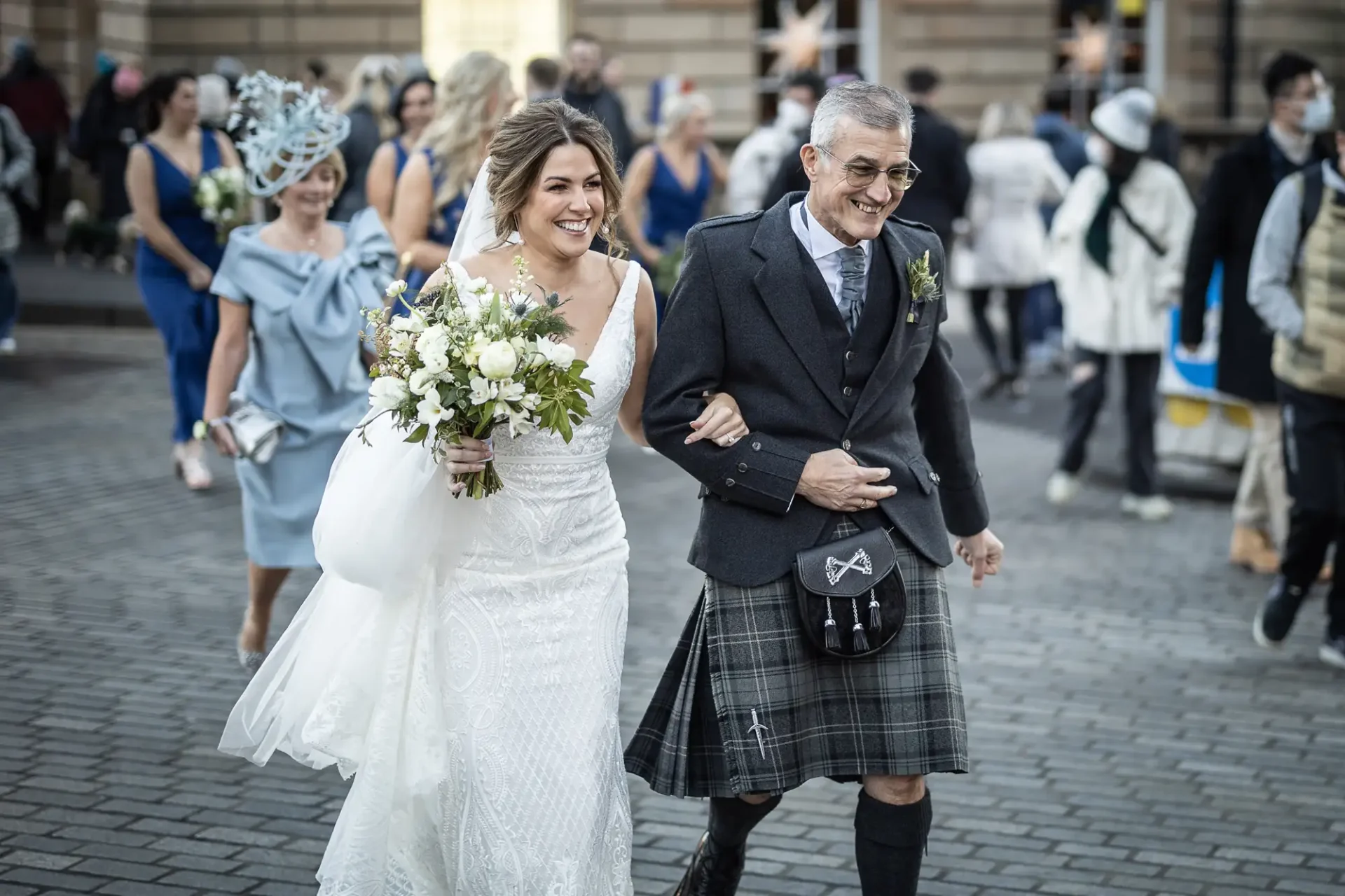 A bride in a white dress and a groom in a kilt and jacket, smiling and walking together on a city street with onlookers in the background.