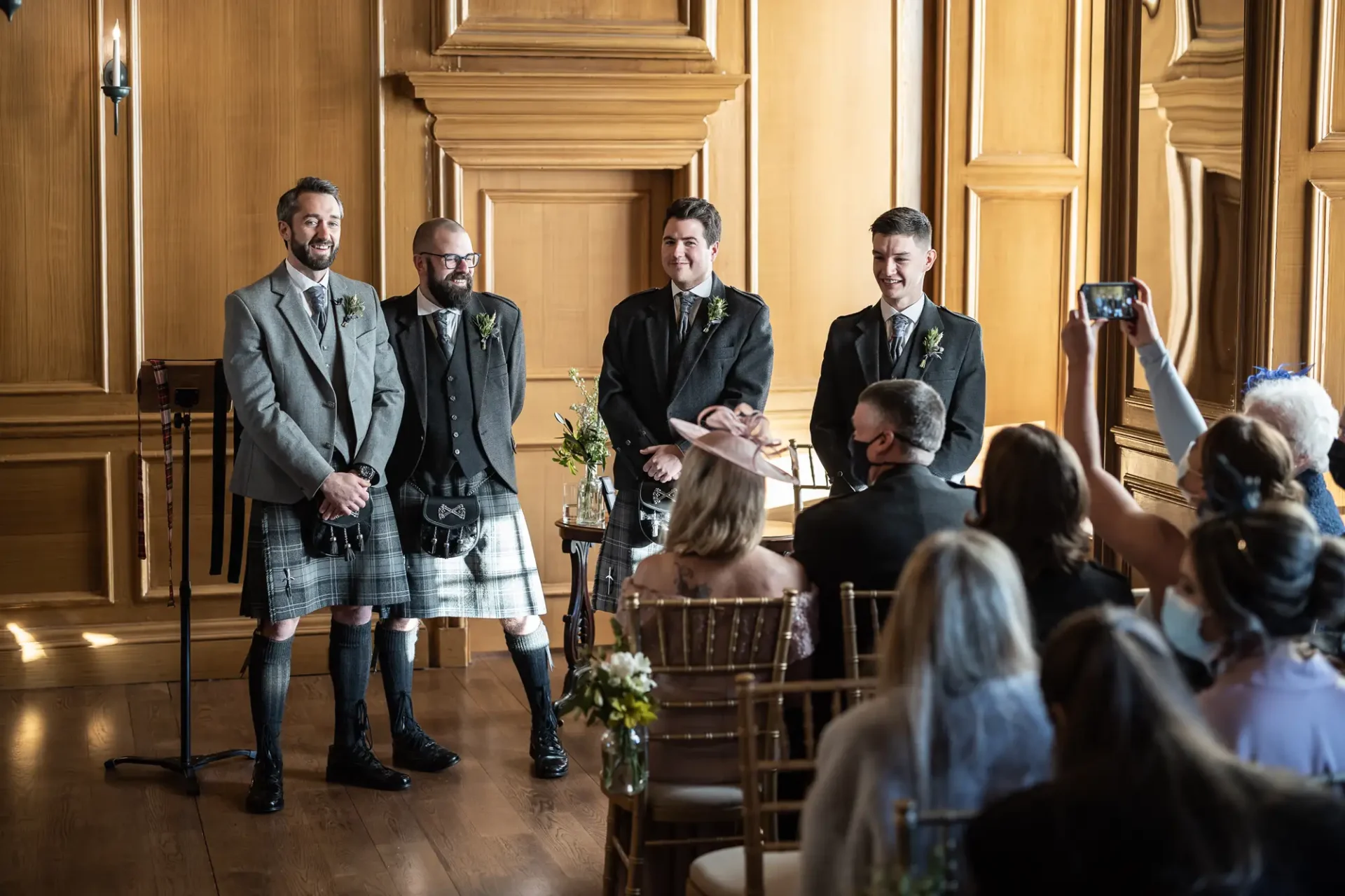 Four men in kilts and jackets stand at the front of a room during a wedding ceremony, with guests seated and watching.