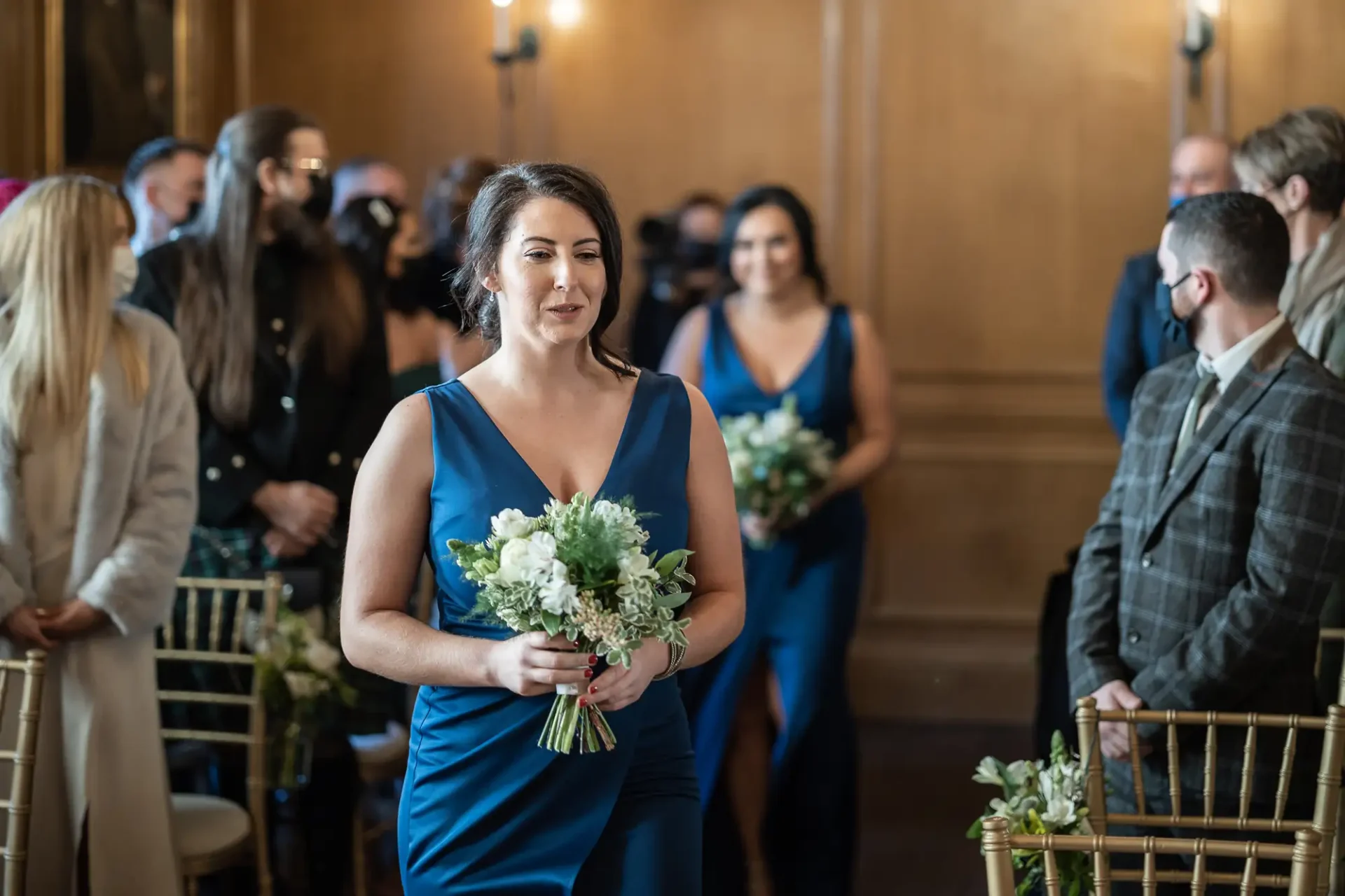 A woman in a blue dress walks down the aisle holding a bouquet, with wedding guests watching from their seats.