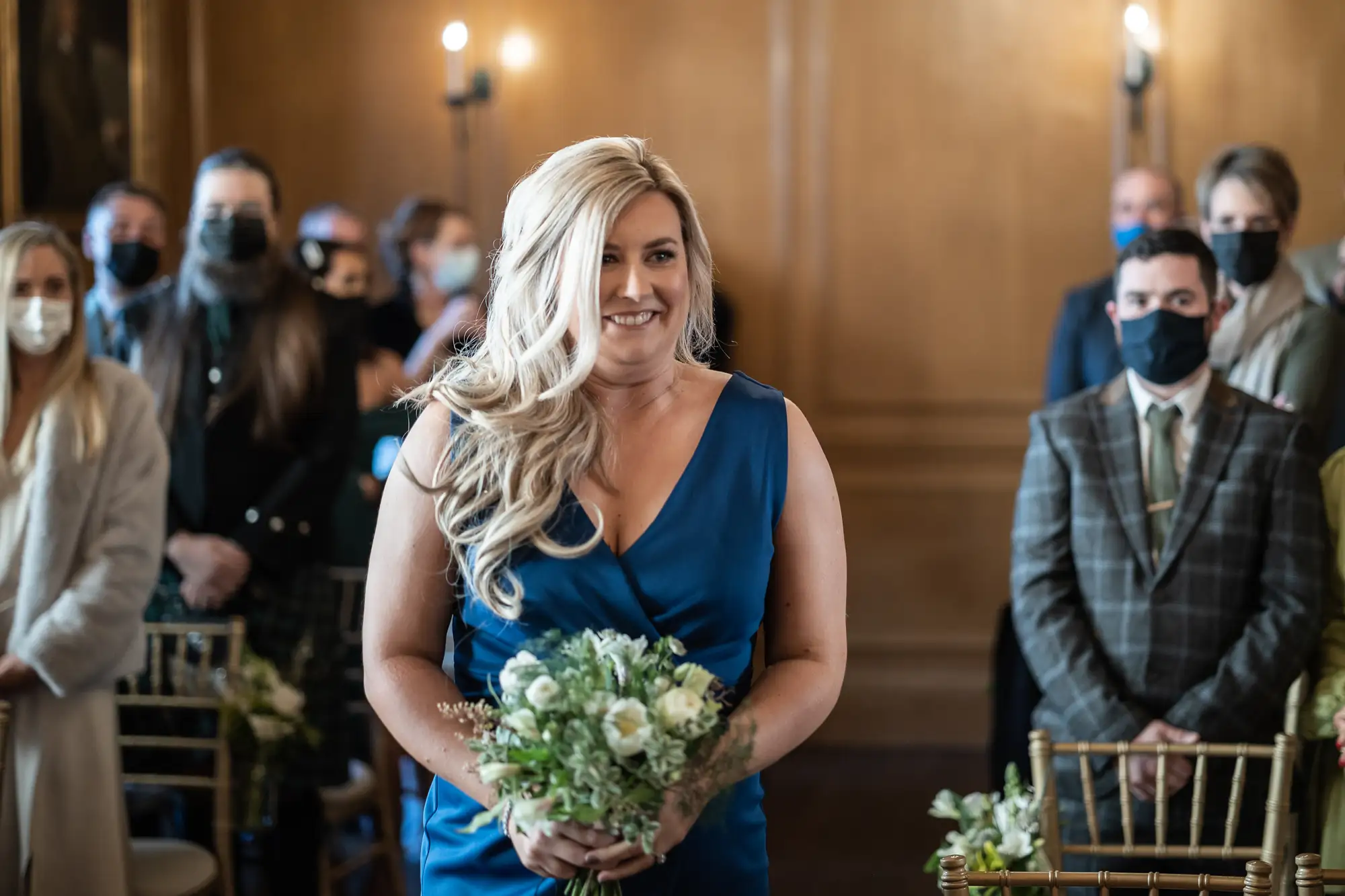 A smiling woman in a blue dress holds a bouquet at a wedding, with guests wearing masks watching her.