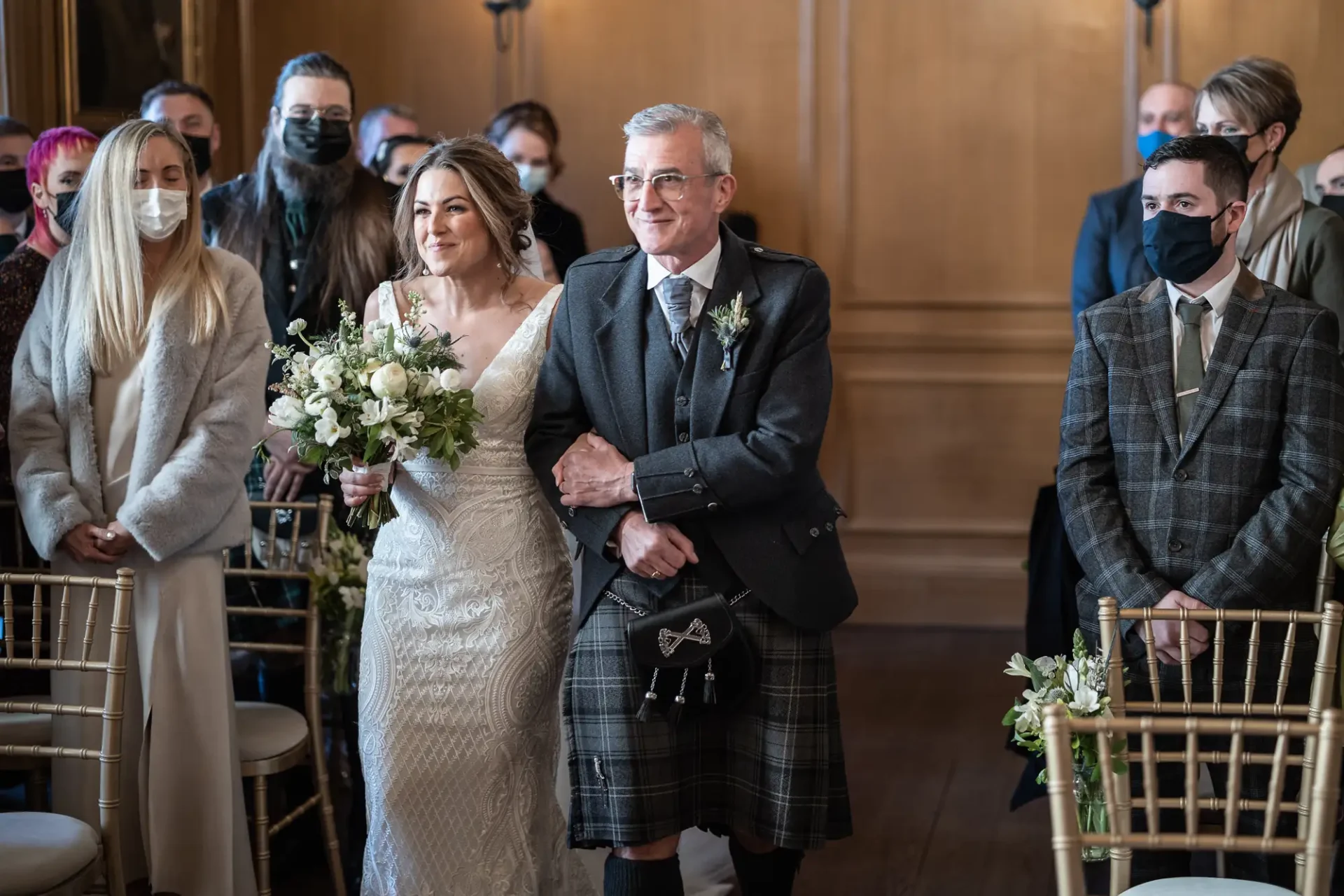 Bride in a lace dress walking down the aisle with an older man in a kilt, surrounded by guests wearing masks.
