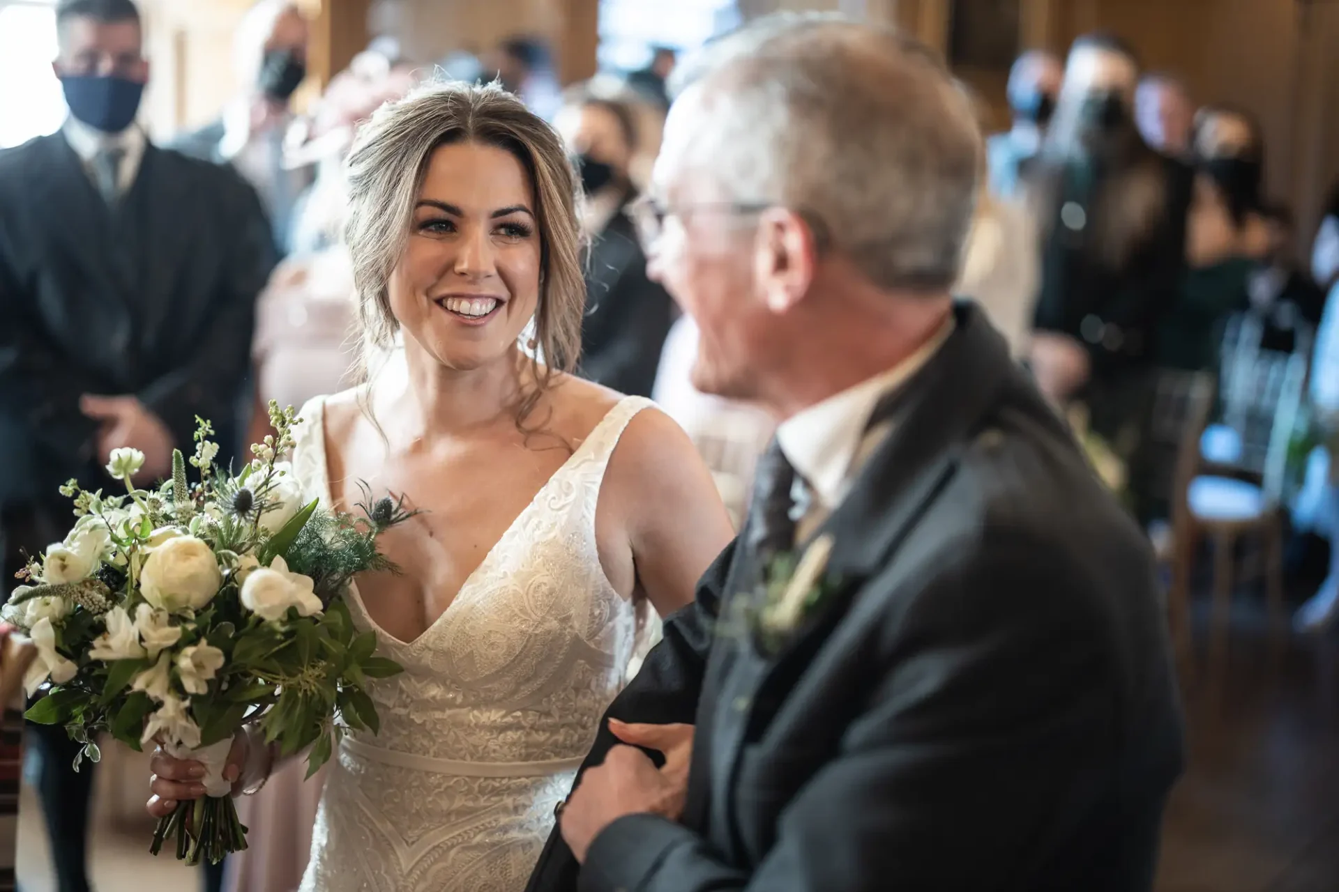 A bride holding a bouquet smiling at an older man in a suit while walking down the aisle, with guests wearing masks in the background.