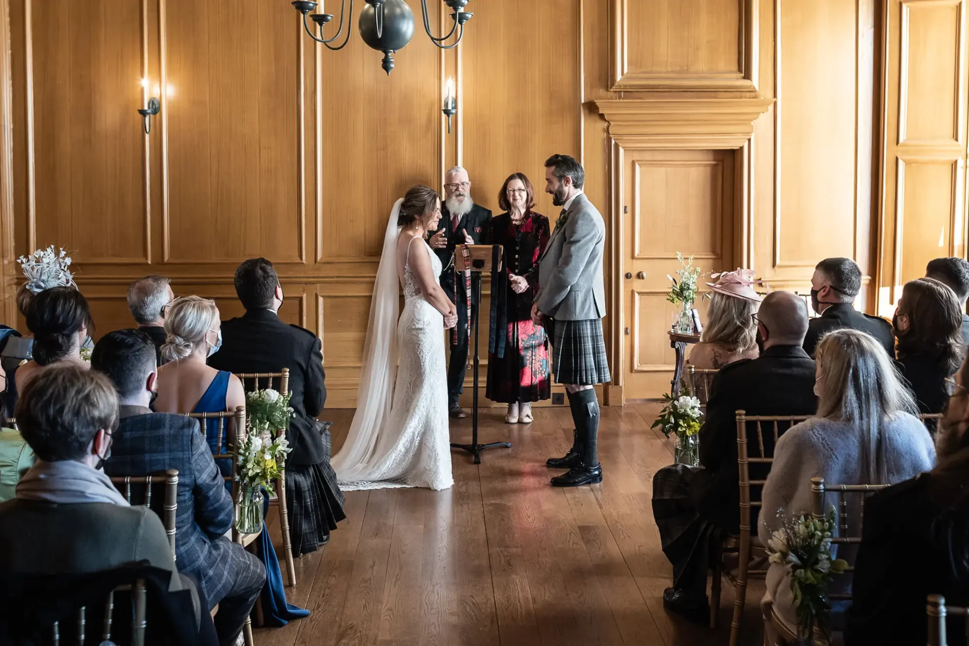 A bride and groom exchange vows at a wedding ceremony in a wood-paneled room with guests seated around them.
