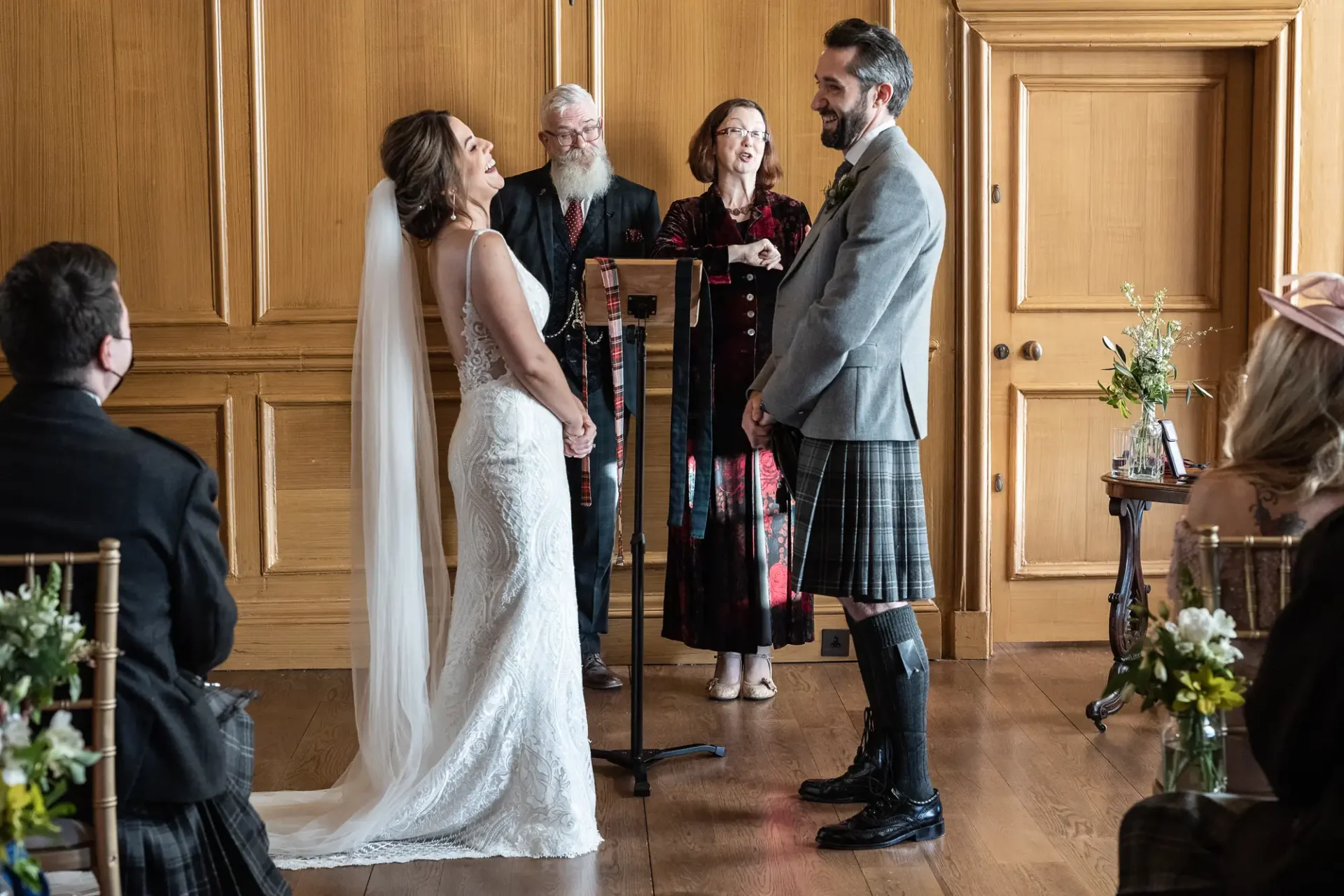 Bride and groom smiling at each other during their wedding ceremony, with an officiant and attendees in a wood-paneled room. the groom wears a kilt.