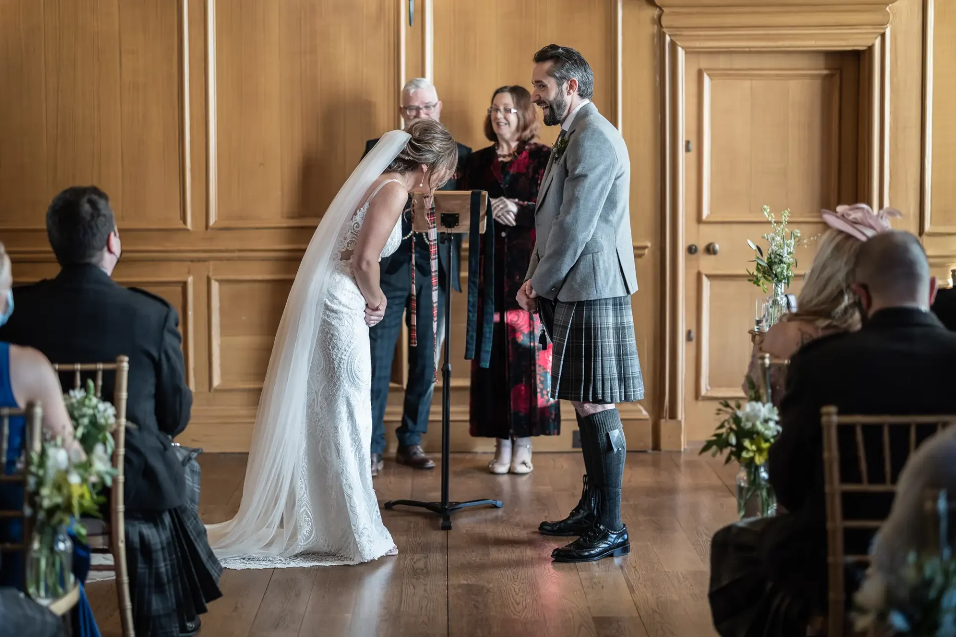 Bride and groom performing a handfasting ceremony in front of an officiant and guests in a wood-paneled room, groom wearing a kilt.