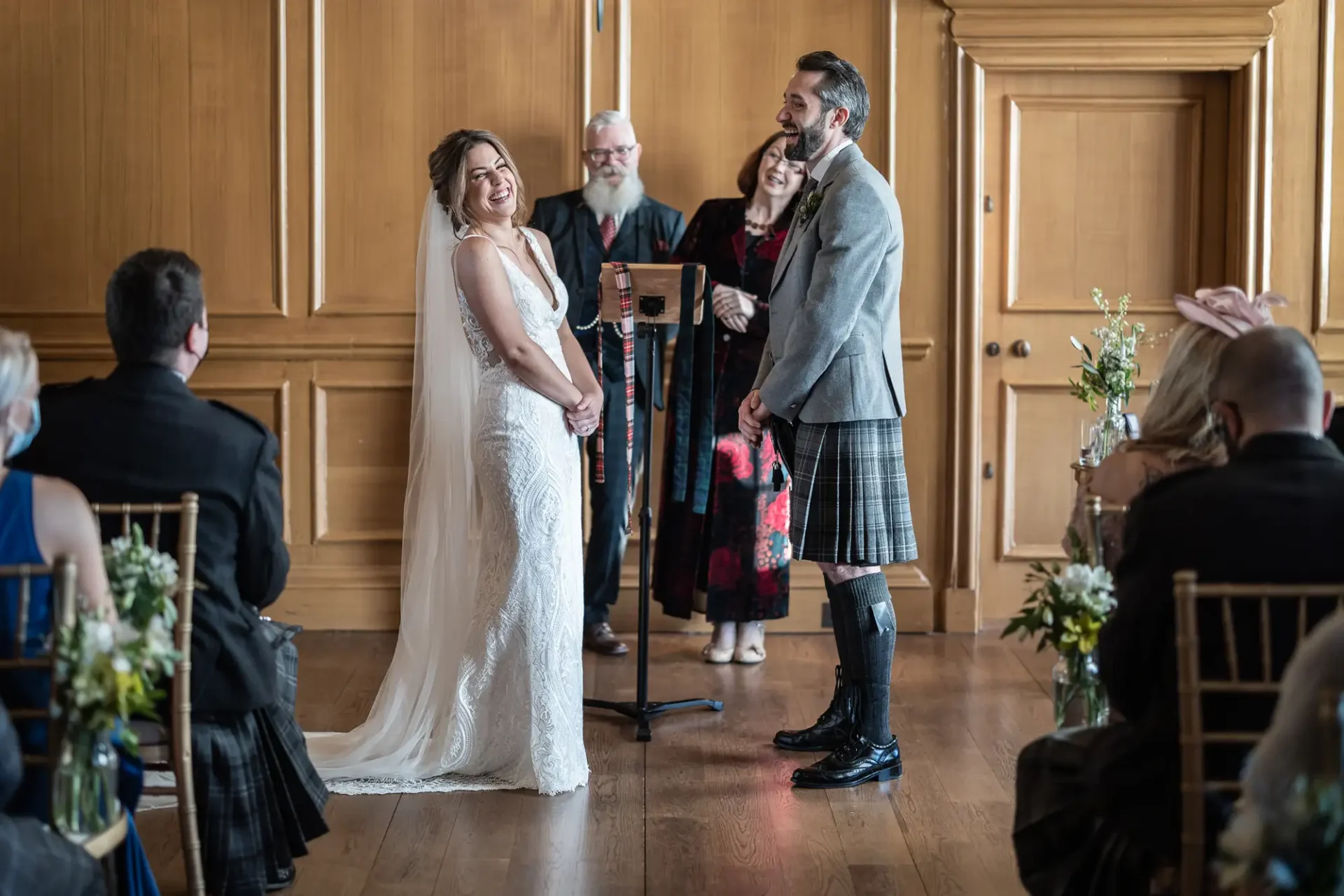 Bride and groom smiling at each other during a wedding ceremony, with the groom wearing a kilt, in a room with wooden paneling and seated guests.