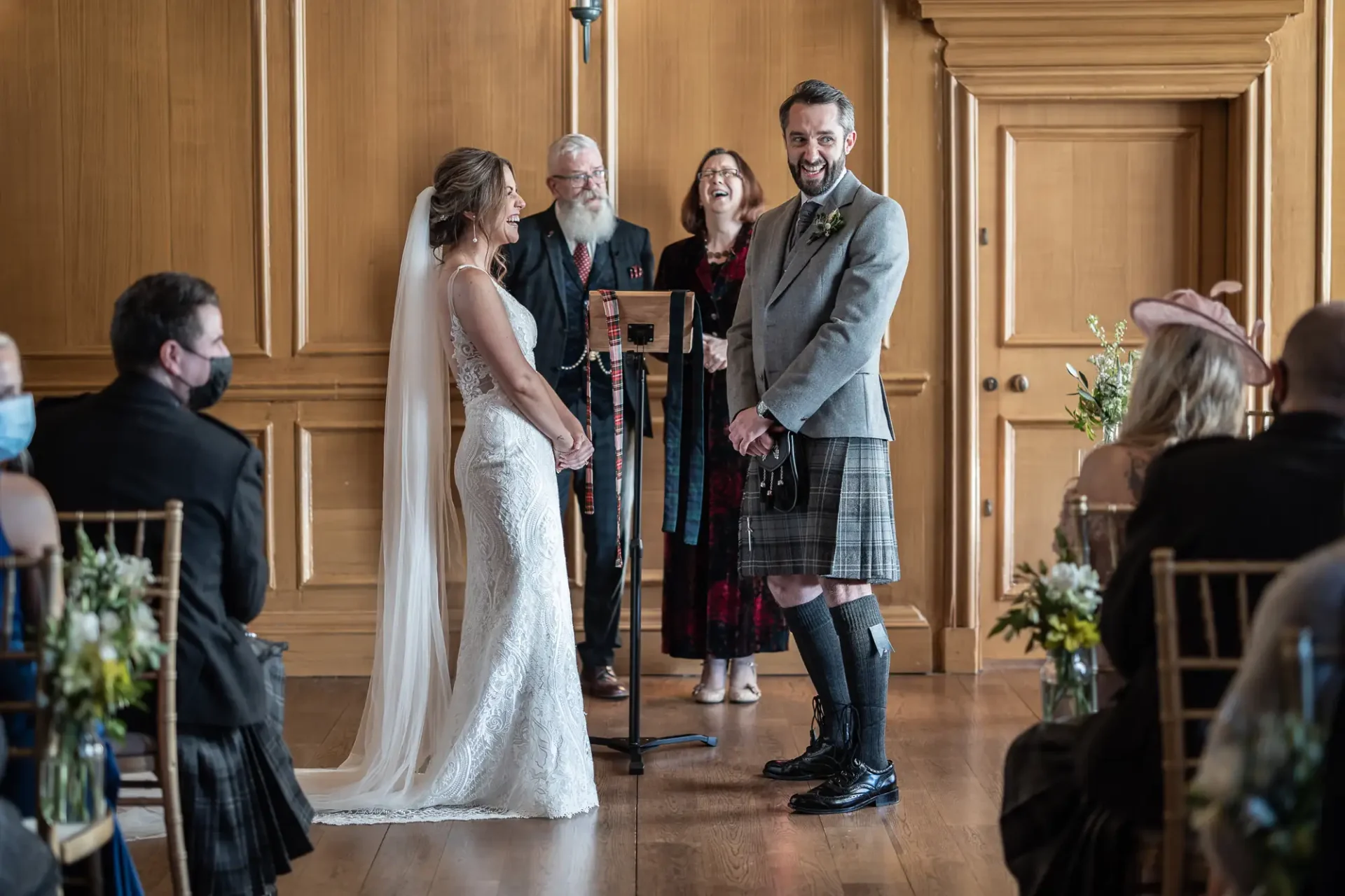 A bride and groom exchanging vows at a wedding ceremony, attended by guests and an officiant in a wood-paneled room. the groom wears a kilt.