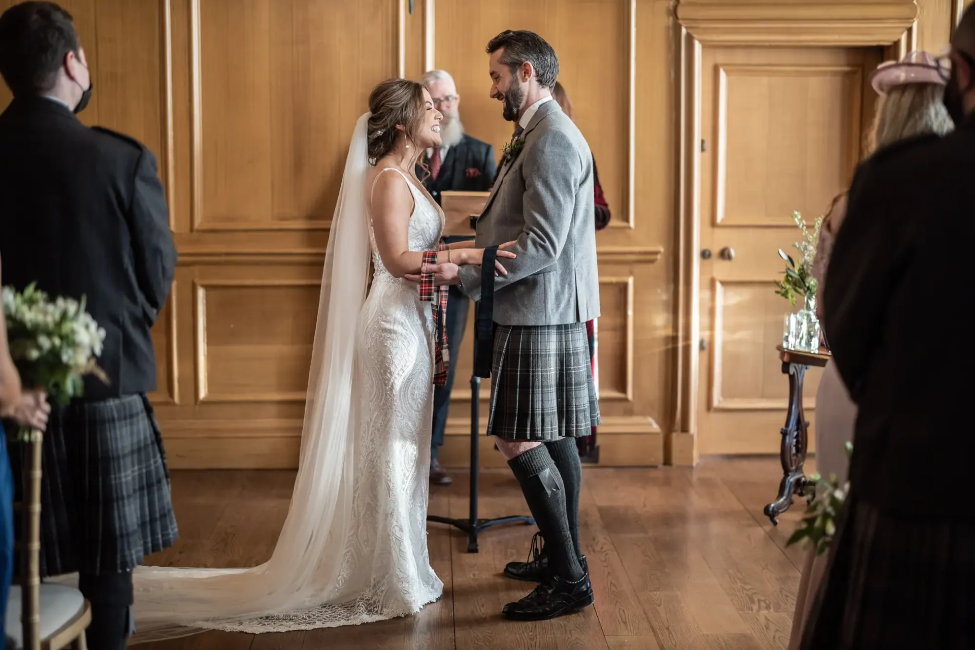 A bride and groom exchange vows, the groom wearing a kilt; guests are visible in an elegantly paneled room.