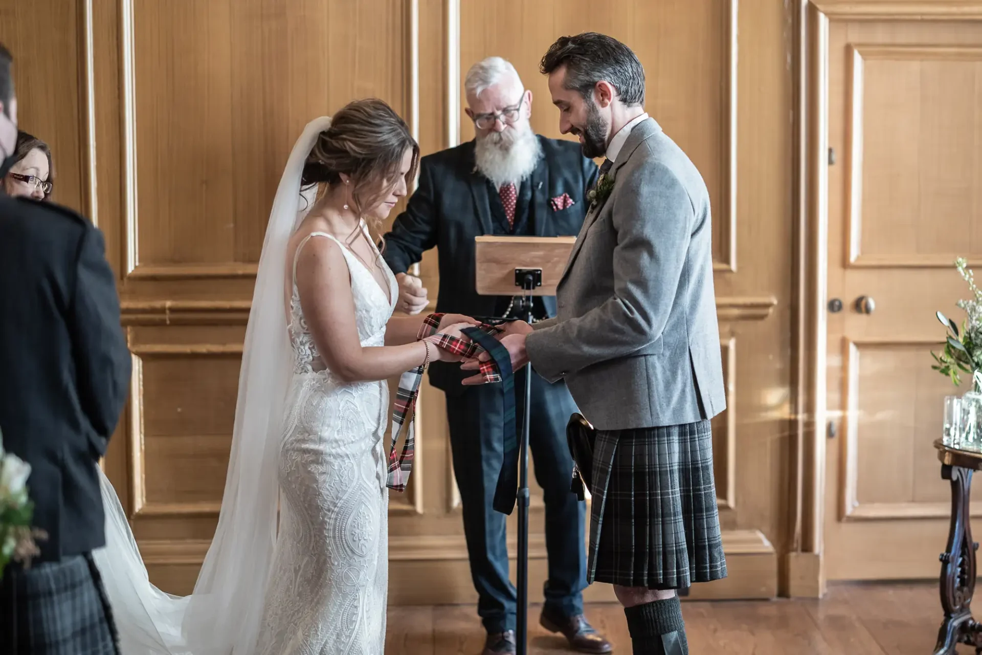 Bride and groom, dressed in a lace wedding gown and kilt, participate in a handfasting ceremony officiated by a bearded man, in a wood-paneled room.