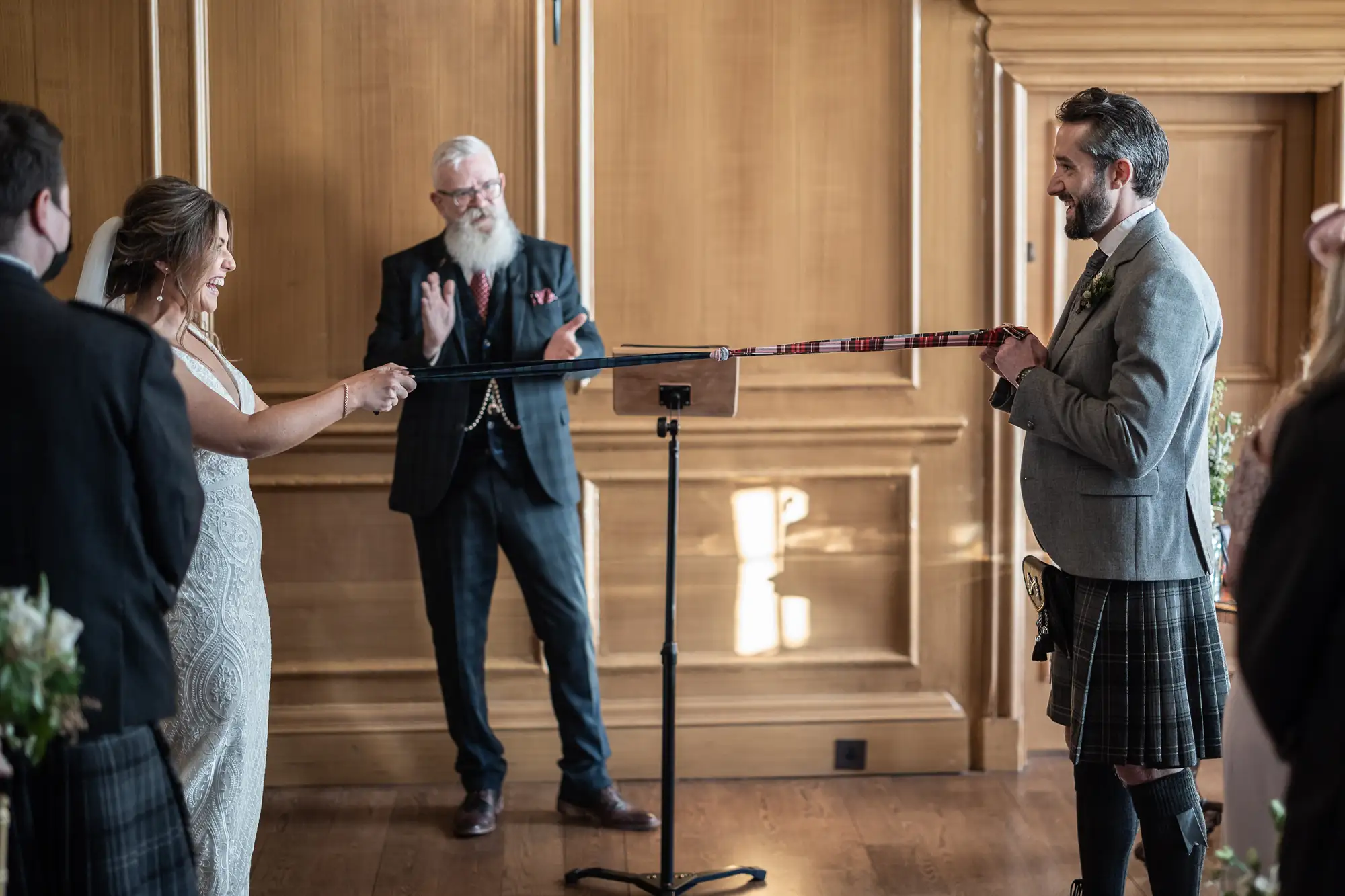 Bride and groom holding a pole at their wedding ceremony, with an officiant speaking in the background in a wood-paneled room.