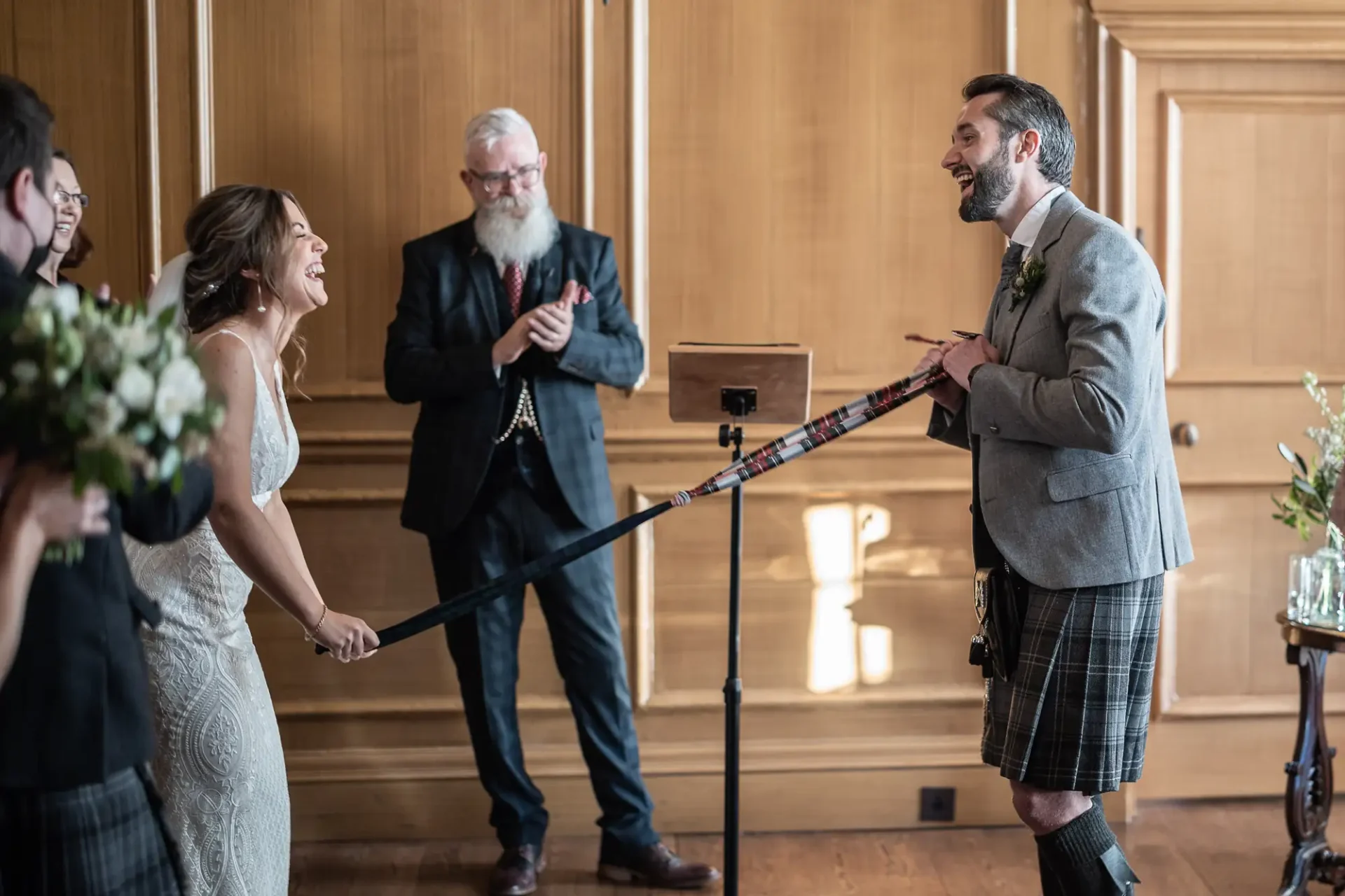 Bride and groom laugh joyously during a wedding ceremony led by an officiant in a wood-paneled room, with the groom wearing a traditional kilt.