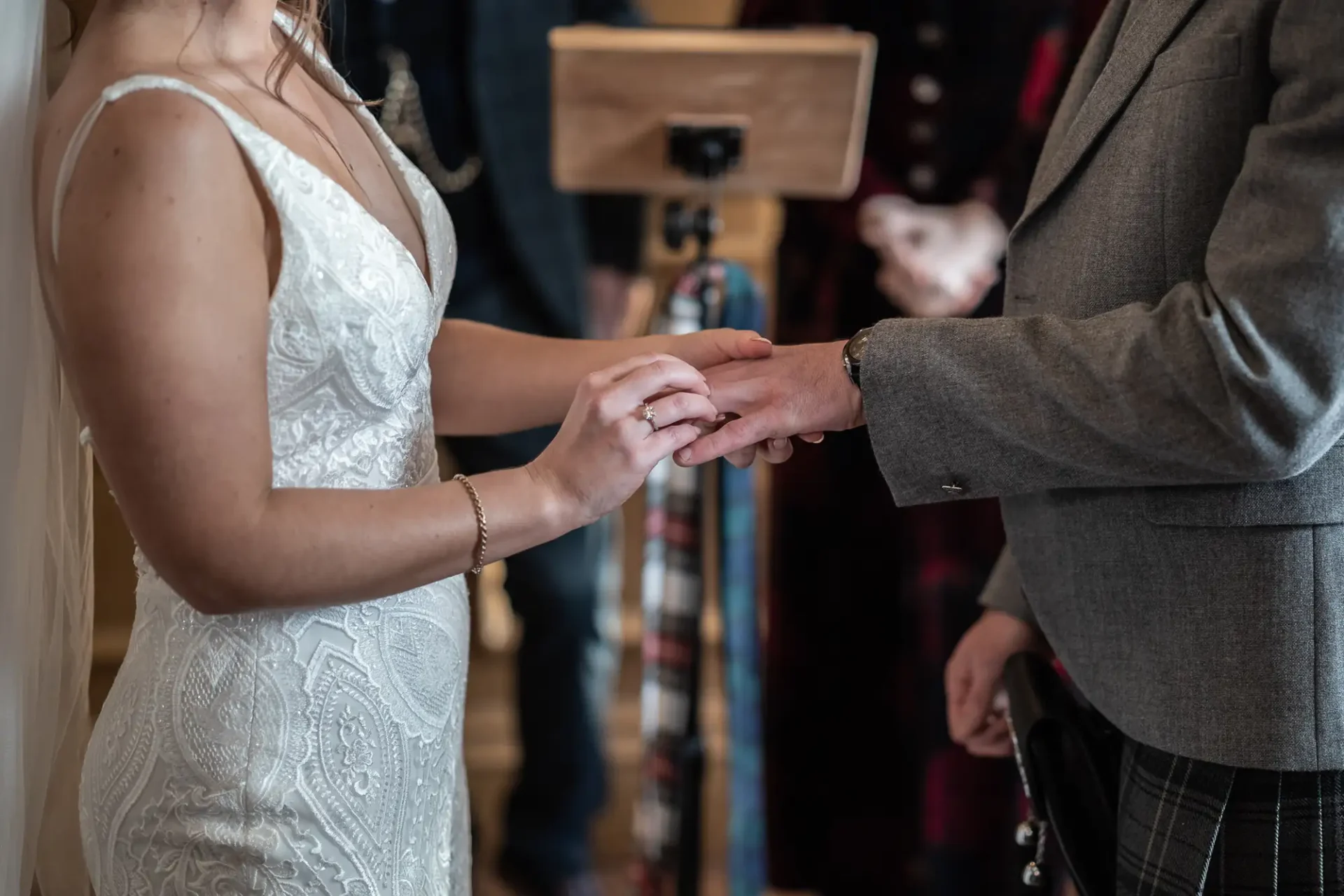 Bride in a lace dress exchanges rings with groom in a tartan kilt during a wedding ceremony.