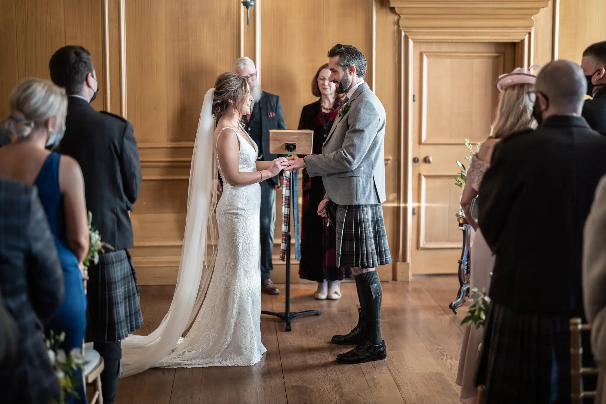 A bride in a white dress and a groom in a kilt exchanging vows in a wood-paneled room, surrounded by guests.