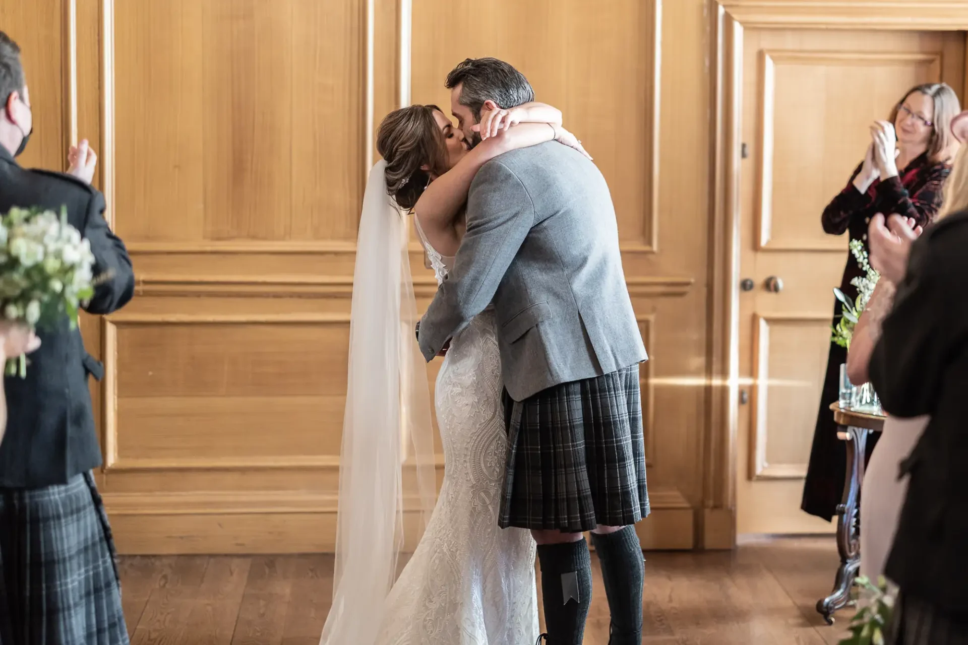 A bride and groom kiss passionately in a wood-paneled room while guests, some in kilts, watch from the sides.