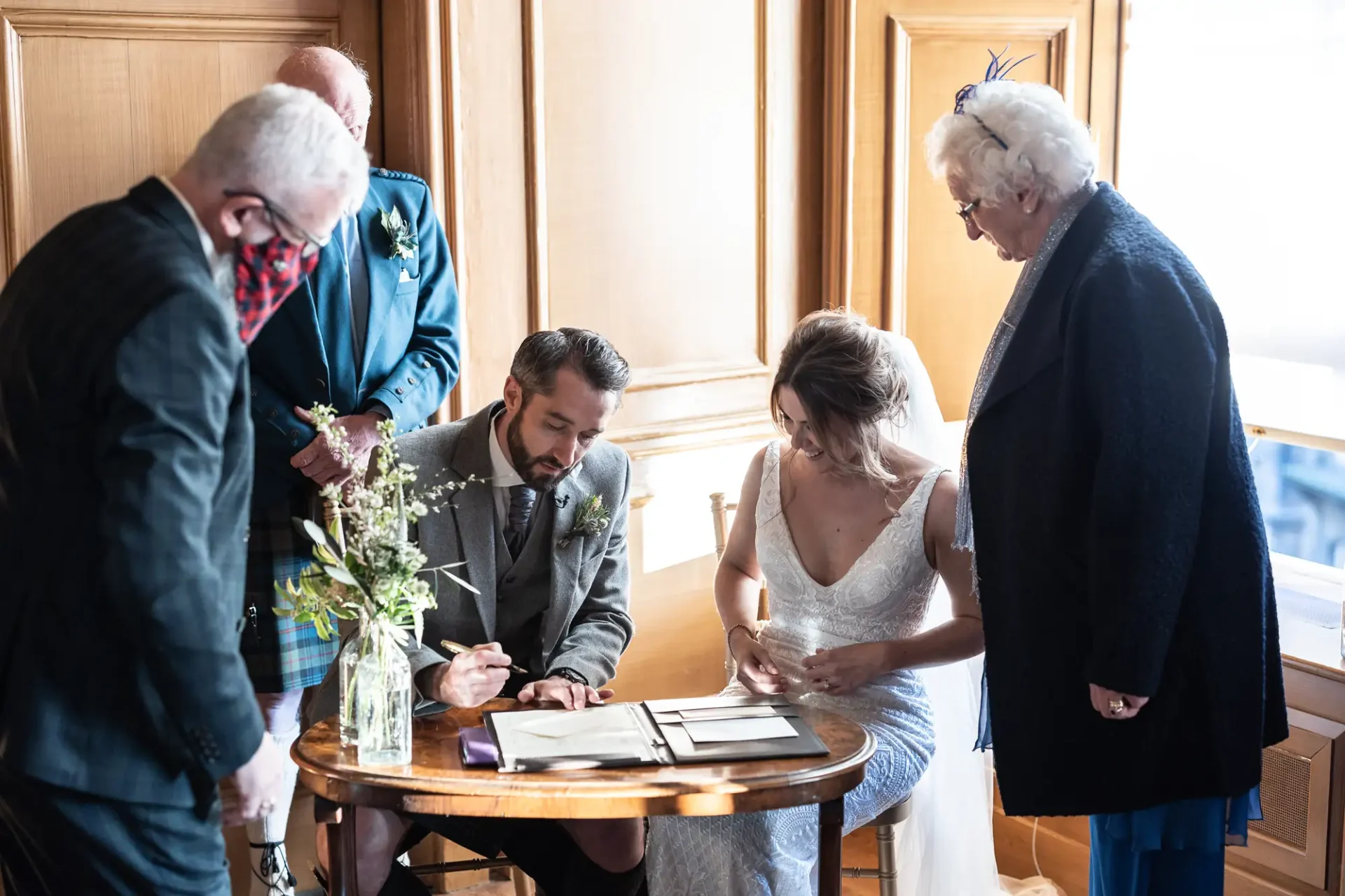 A bride and groom sign a registry at a small table, watched by three older guests, in an elegantly lit room.