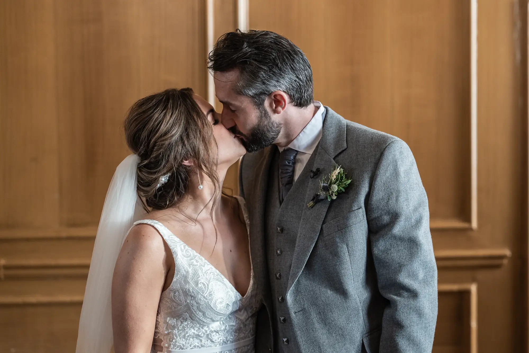 A bride and groom kissing, dressed in wedding attire, inside a room with wooden panels.