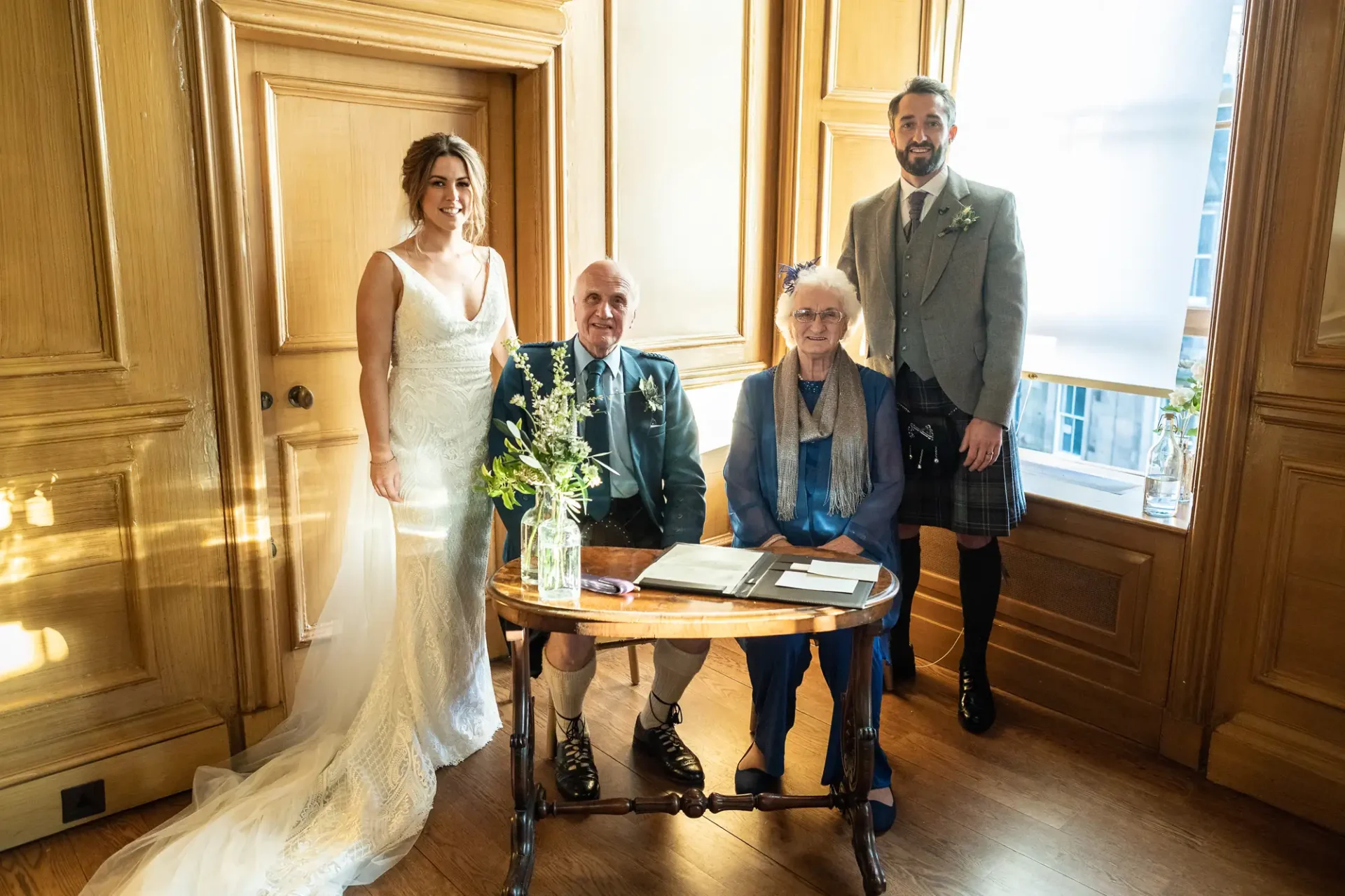 A bride and groom stand beside an elderly couple seated at a table with a floral arrangement in a warmly lit room.