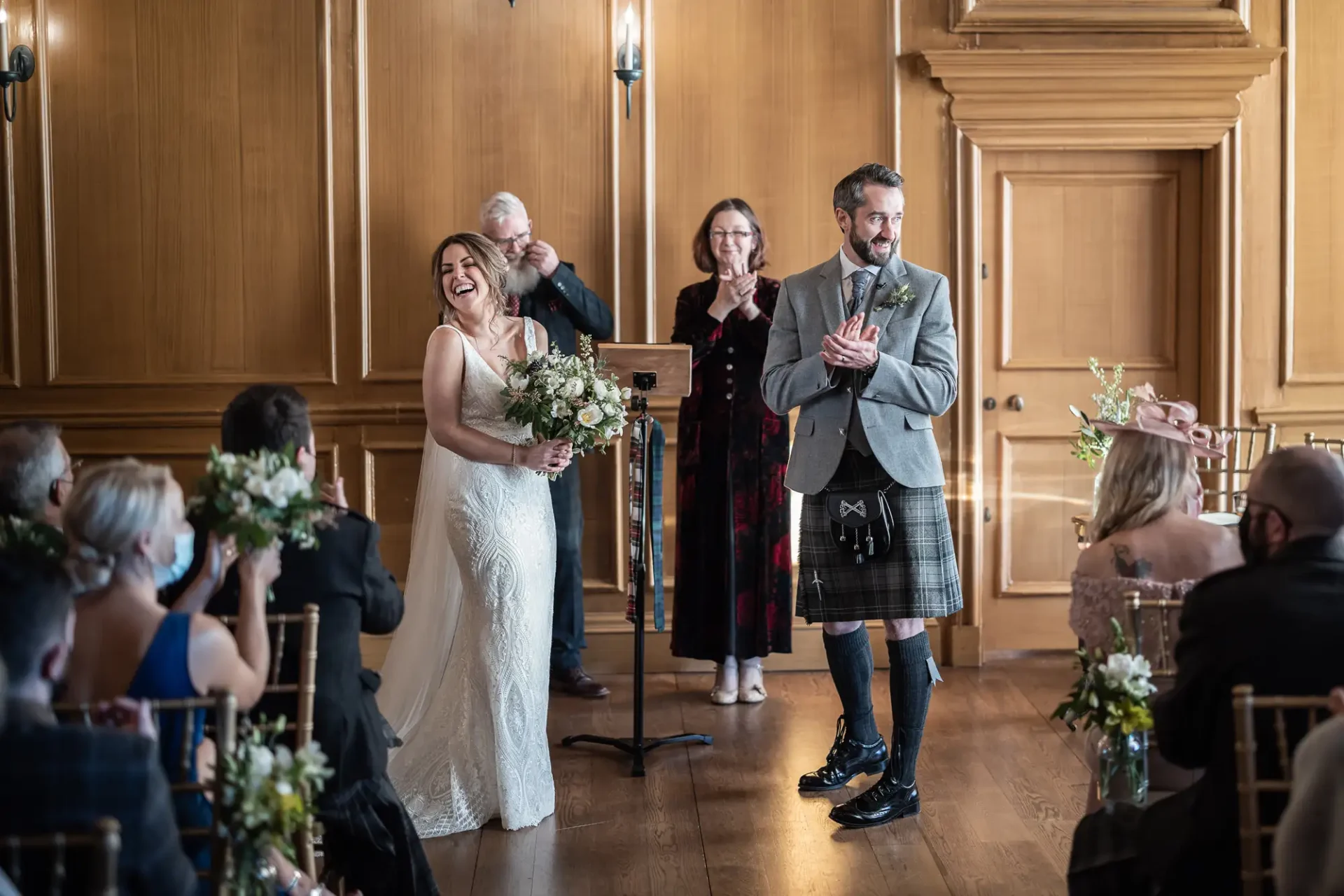 A joyful wedding scene in a grand hall: a bride and groom, the latter in a kilt, smiling brightly as they walk down the aisle, with guests applauding around them.