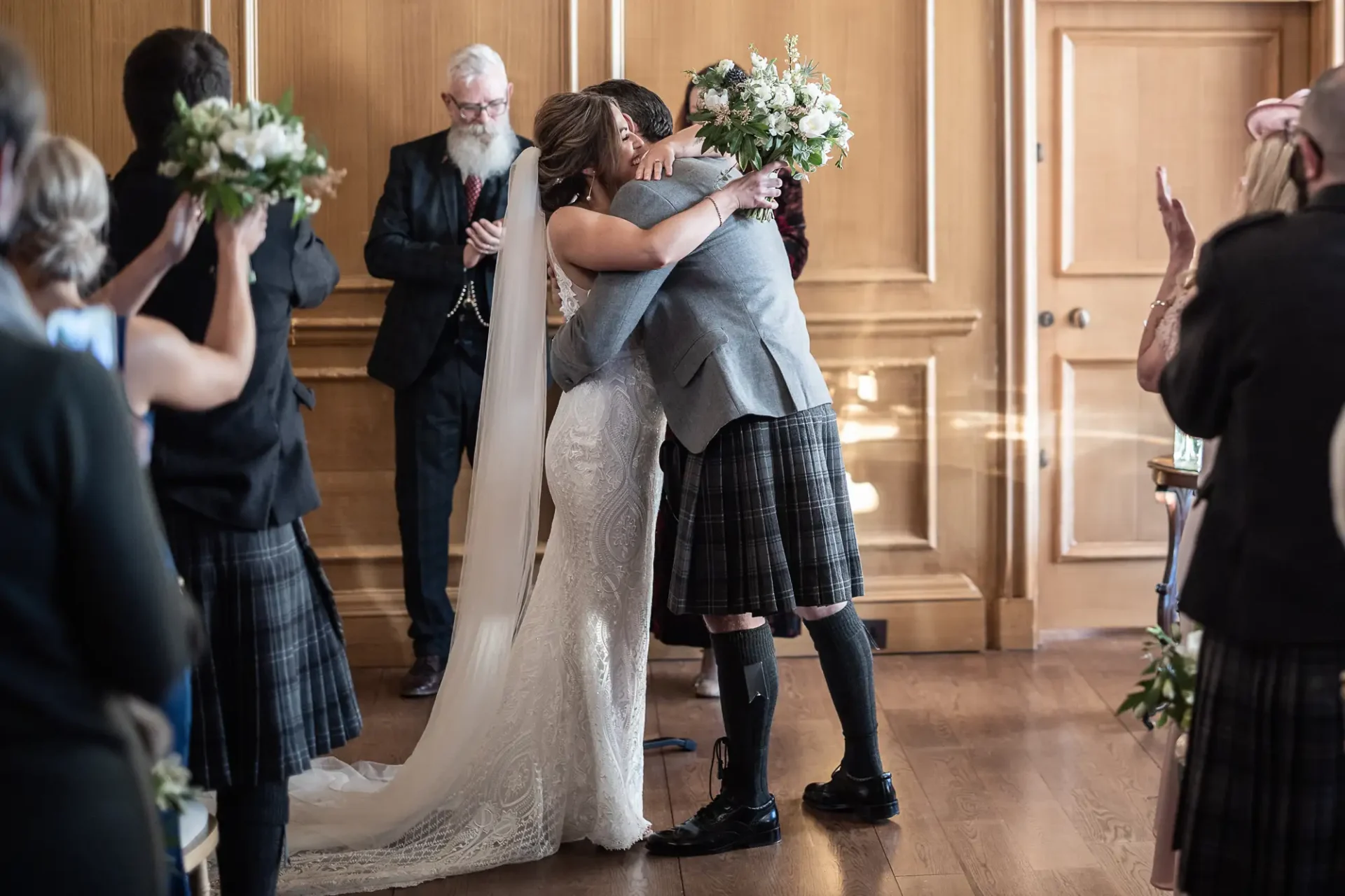 A bride and groom embrace at their wedding ceremony while guests applaud, with the groom wearing a kilt and the bride in a lace gown.