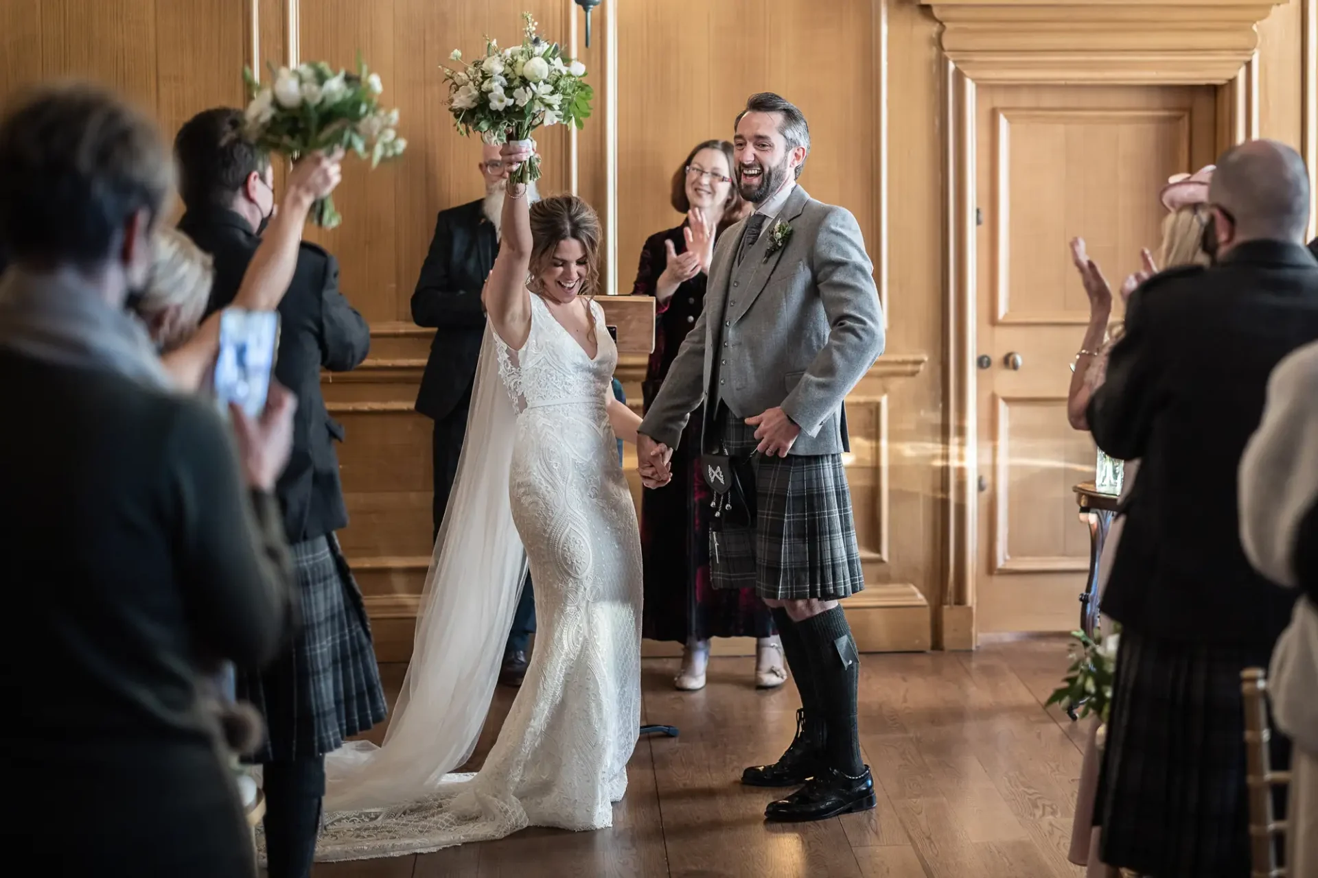 A joyful bride and groom walk down the aisle while guests applaud, the groom in a kilt and the bride holding a bouquet, both smiling broadly.