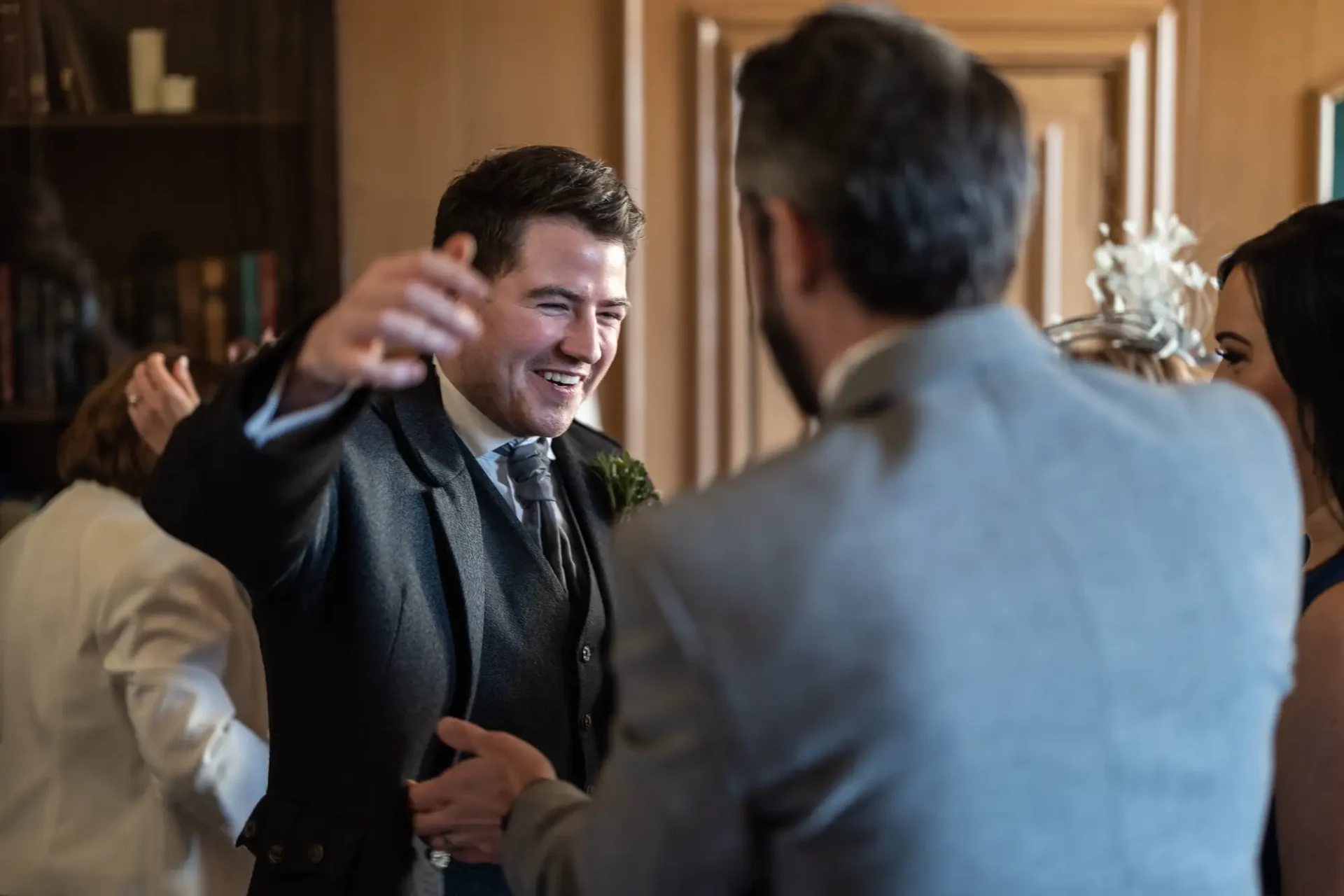 A man in a suit smiles and gestures while talking to another man at a social gathering.