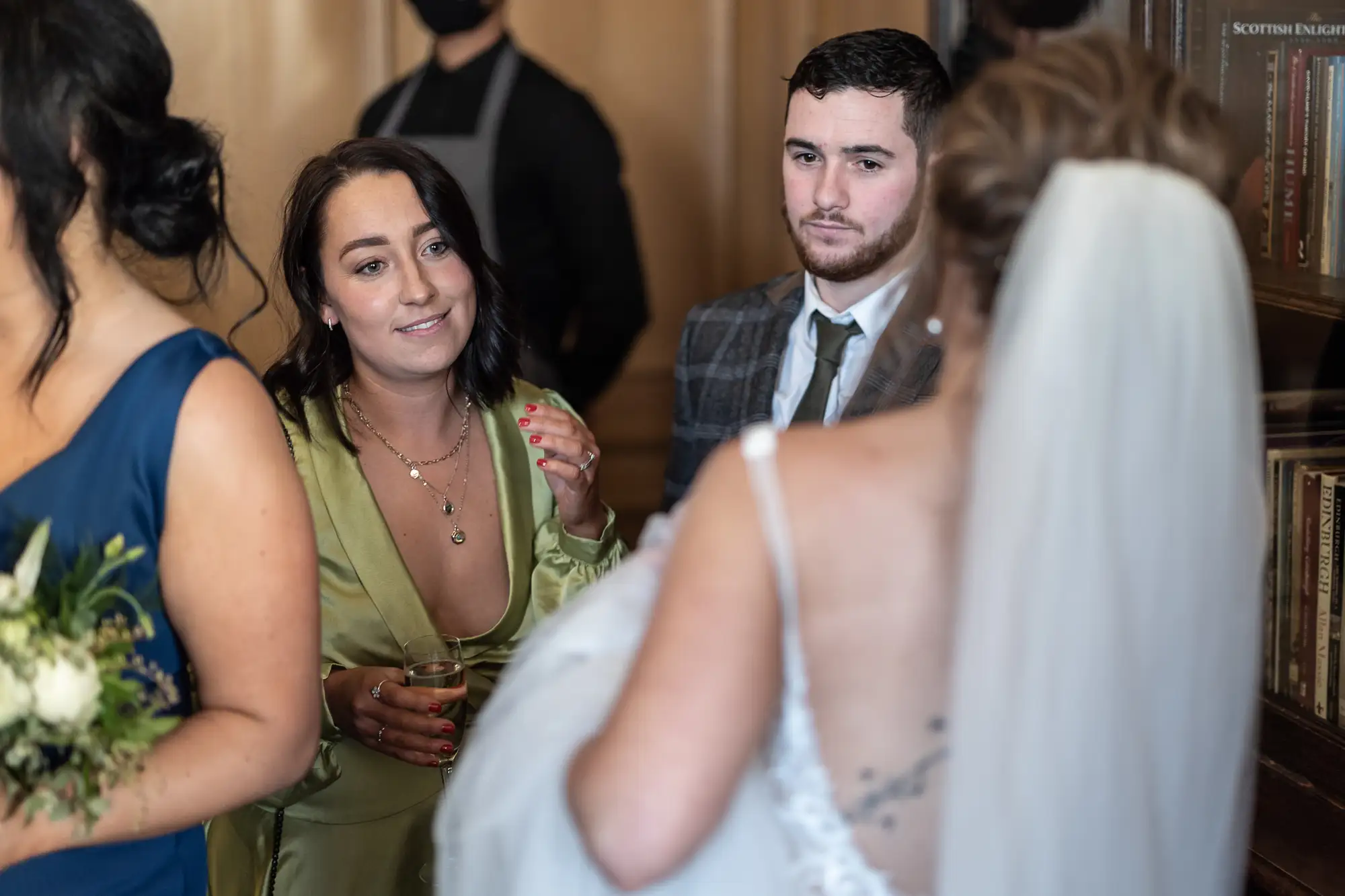 A bride in a white gown listens attentively to a woman in a green dress, with a man in a gray suit between them, in a room with books.