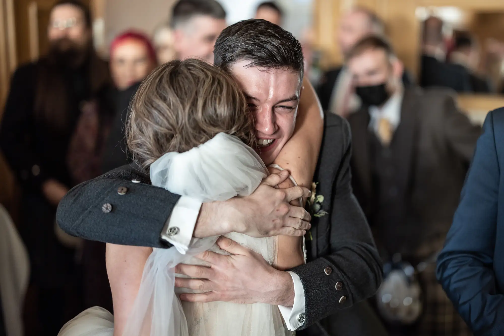 A joyful groom in a black suit embraces a bride in a white dress at a wedding, surrounded by guests wearing masks.