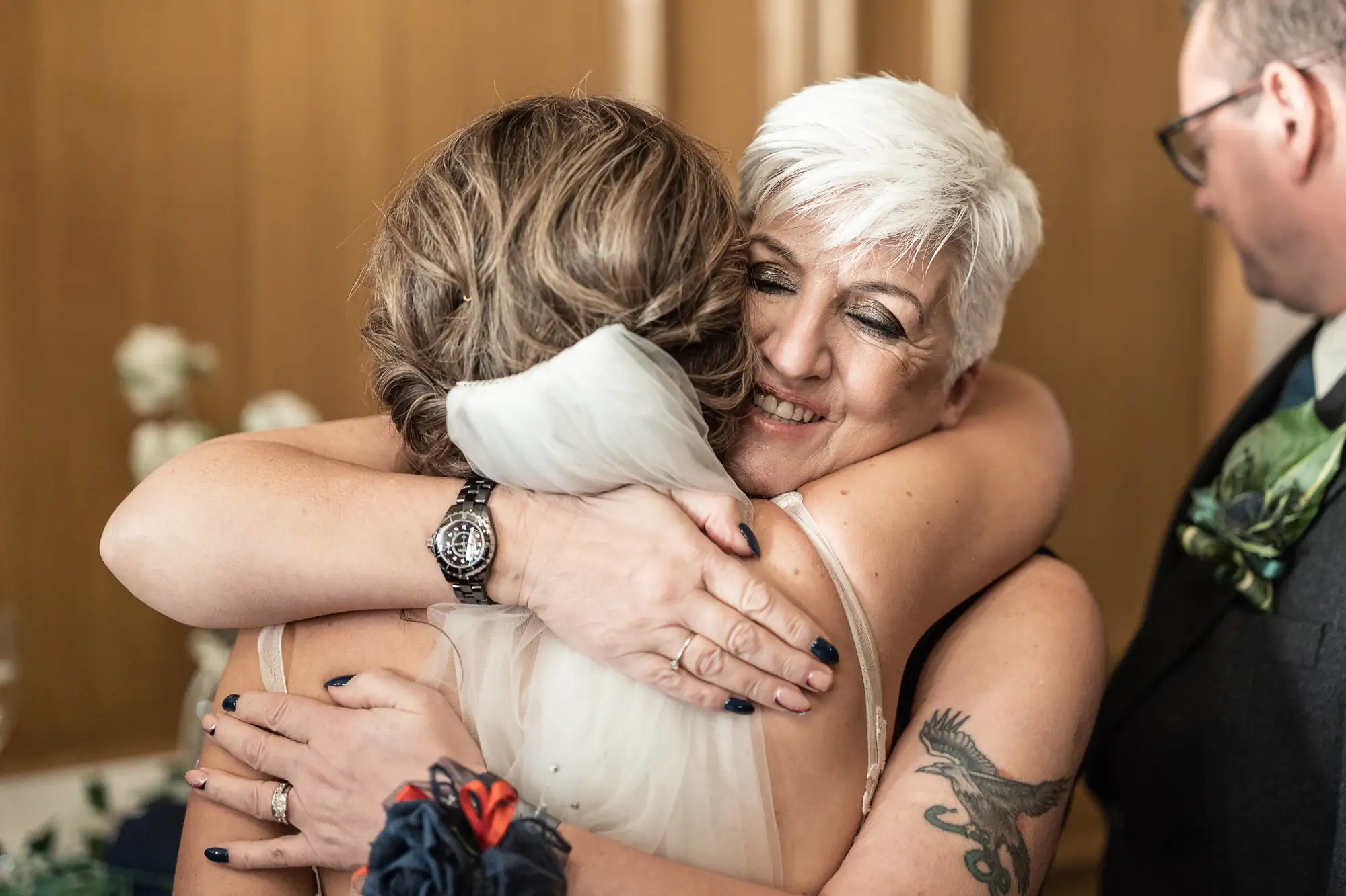 A joyful wedding scene with an older woman embracing a younger bride, both smiling with closed eyes, in a warmly lit room.