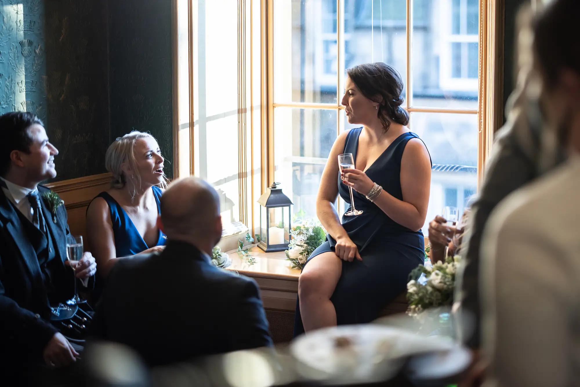 A woman in a navy dress holding a champagne flute, seated at a table with elegantly dressed guests in a warmly lit room.